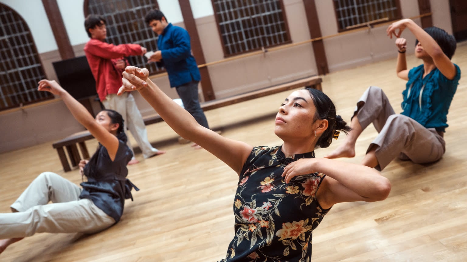 Three dancers dressed in khaki pants and various-colored tops sit on a wooden floor in floaty, reaching poses.