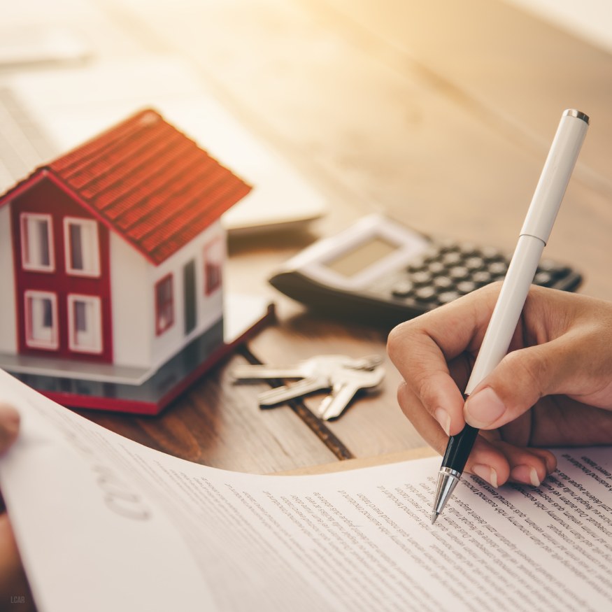hand signing a contract with a pen with keys, a calculator, and a little red roofed house in the background.