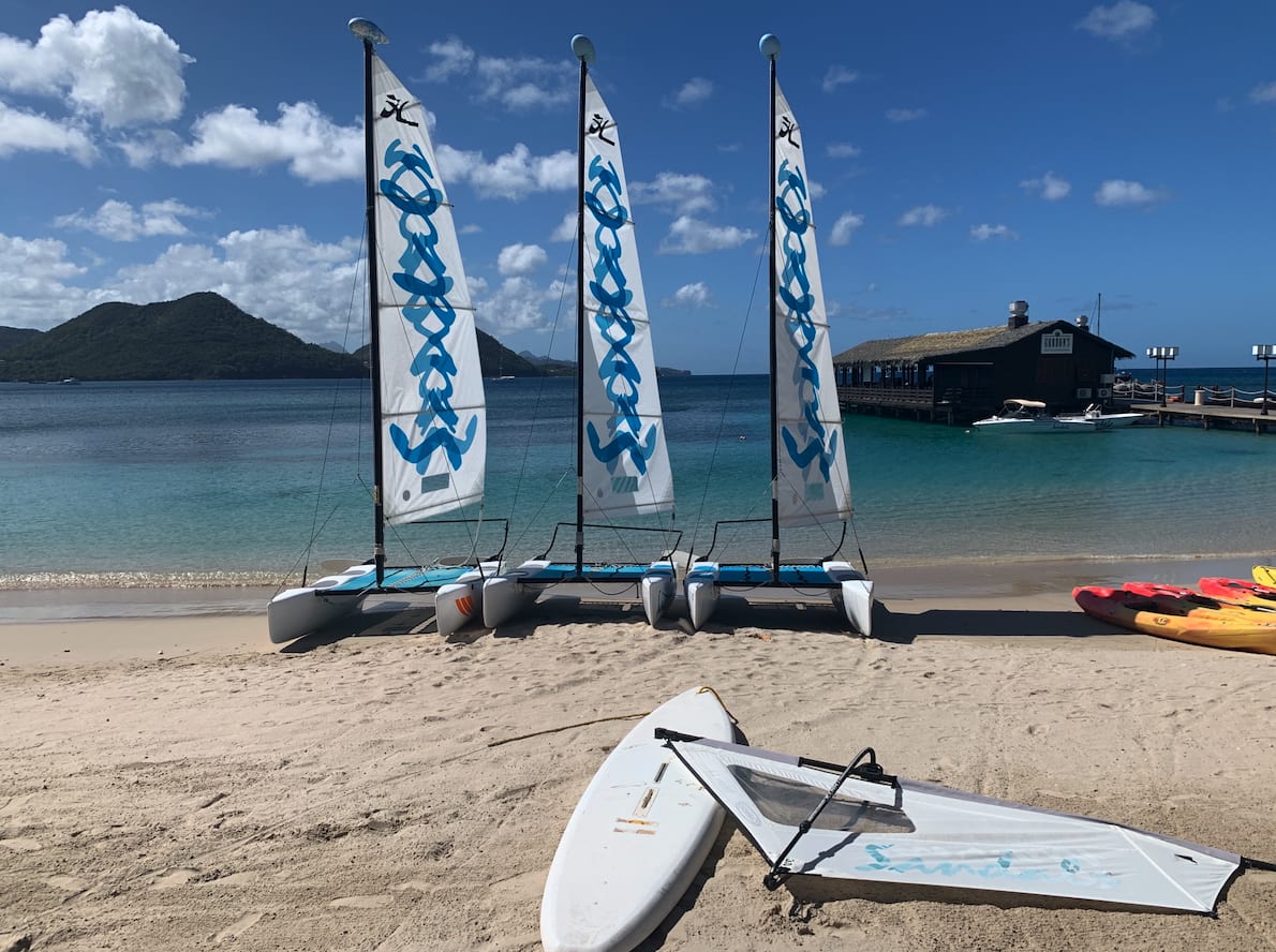 Sailboats, kayaks, and a sailboard on the beach, next to the ocean, with mountains and a restaurant on a pier in the distance