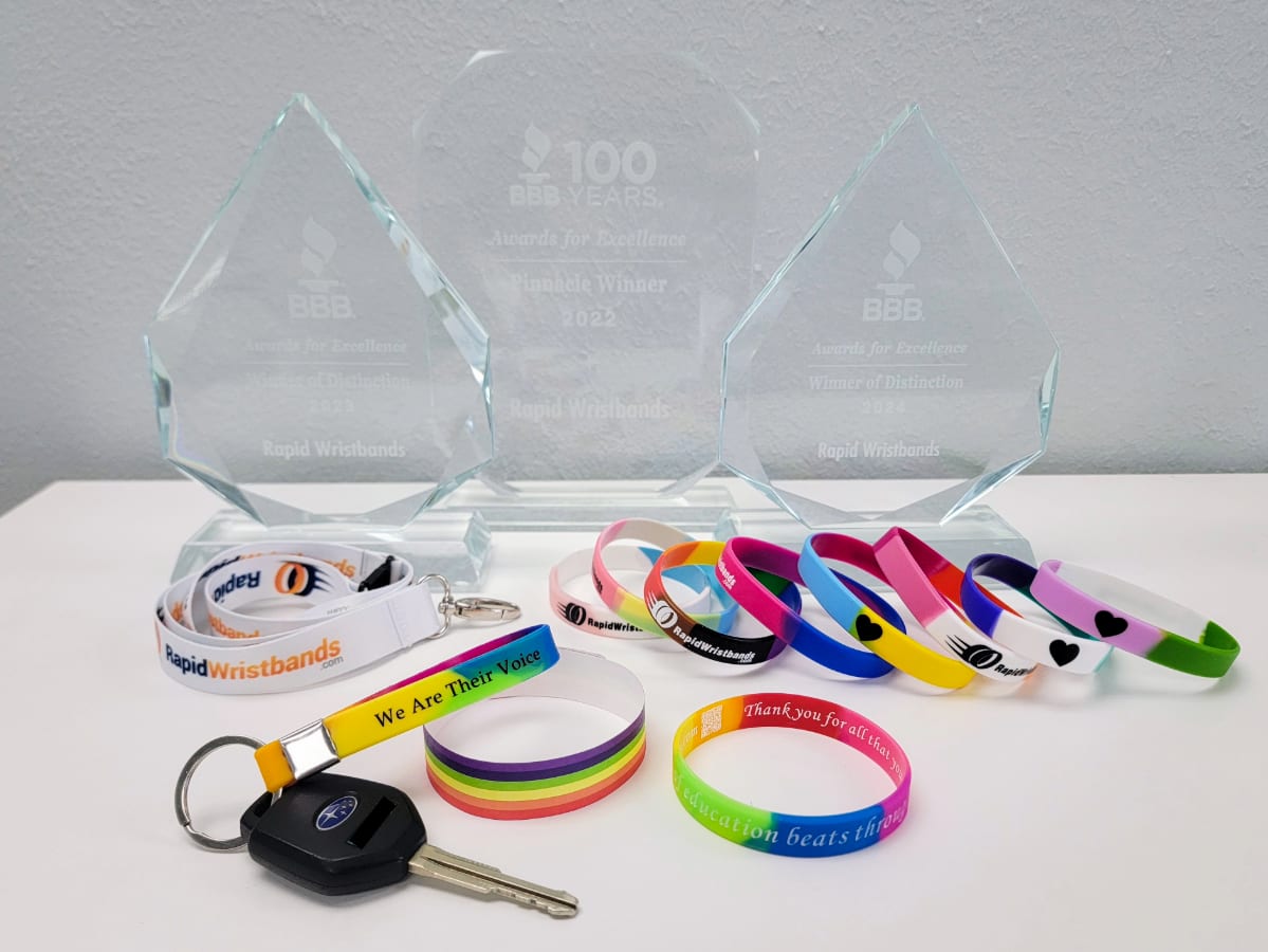 Custom silicone wristbands, cloth wristbands, Tyvek wristbands, keychains, and lanyards in pride flag colors next to trophy.