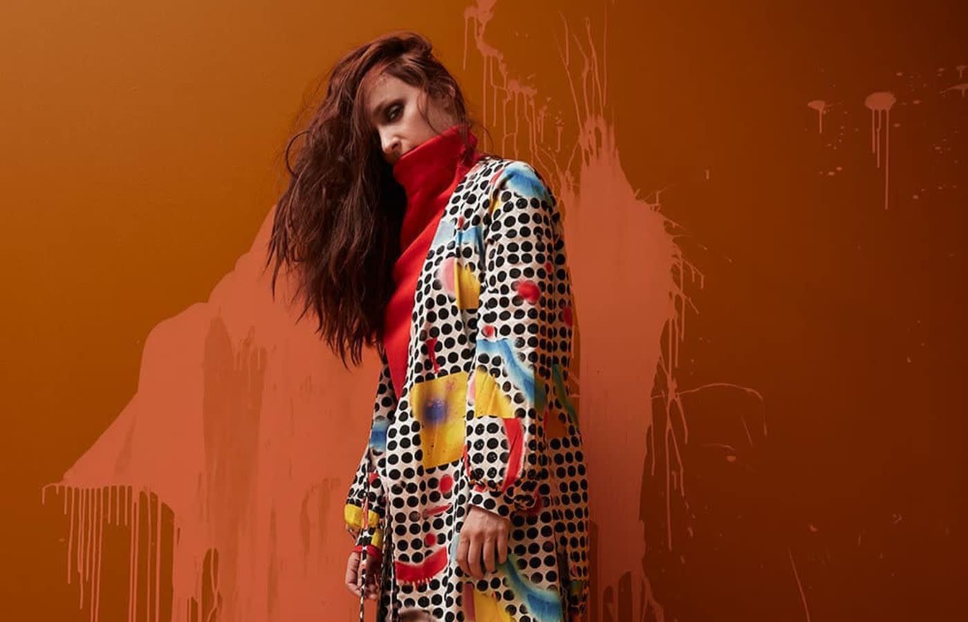 Anneli Kanninen stands in an abstract-patterned outfit against a paint-splattered wall.