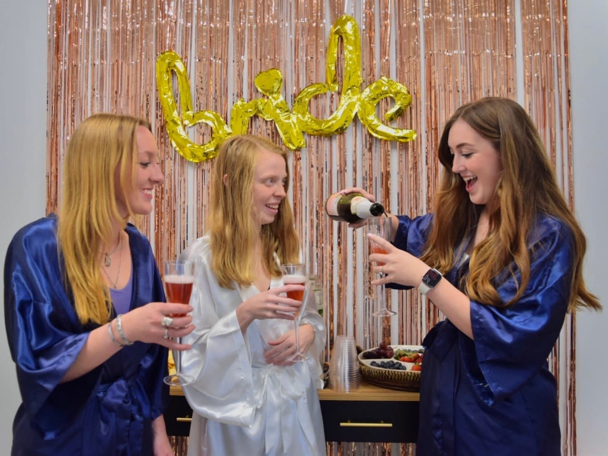 Three women standing in front of a 'Bride' banner, smiling and celebrating as one pours a glass of champagne.