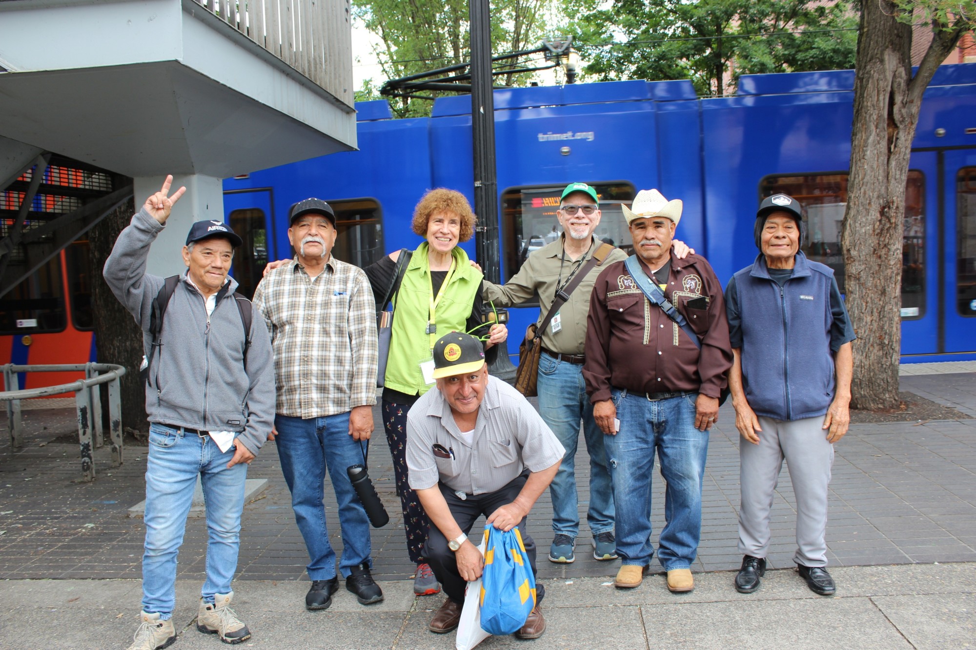 A group of seven people smile and pose together on a sidewalk in front of Trimet Max Light Rail.