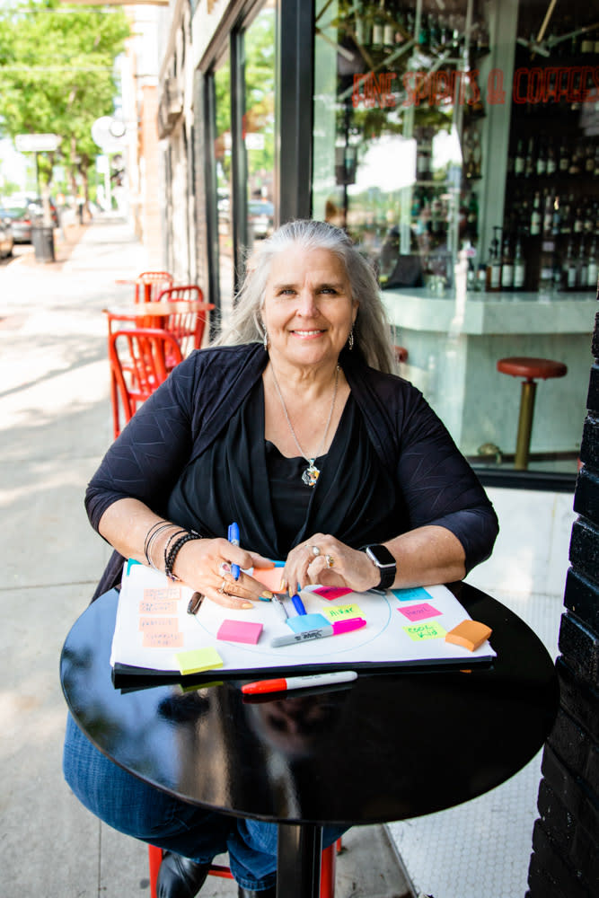Woman sitting at an outdoor café table smiling while holding a marker over a whiteboard covered in colorful sticky notes.