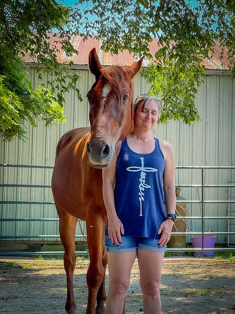 Woman standing with a chestnut horse under a leafy tree, eyes closed and leaning gently into the horse in a shaded ranch sett