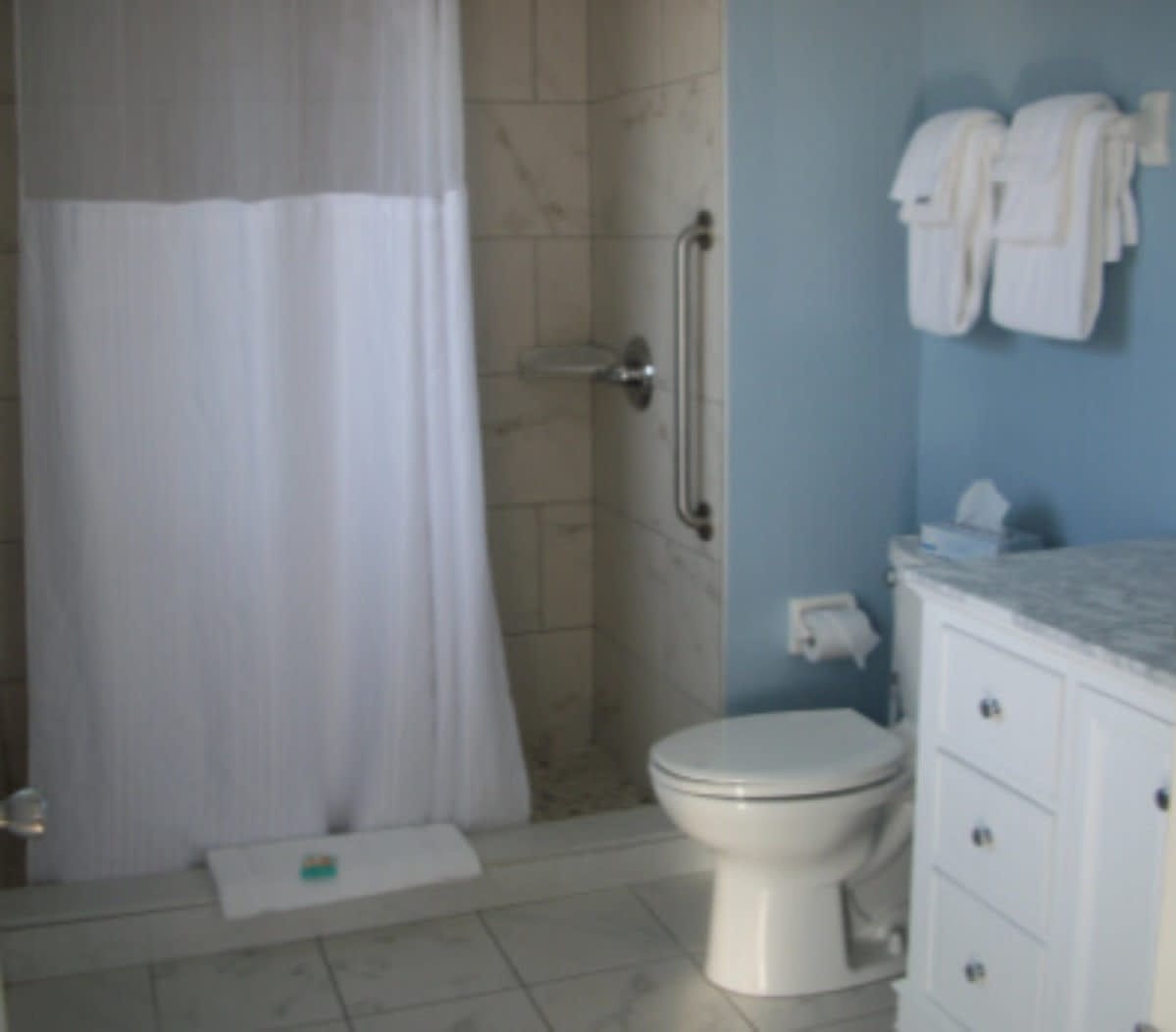 Bathroom with beige tile, walk-in shower with curtain, white vanity, toilet, and towels hanging on wall rack.