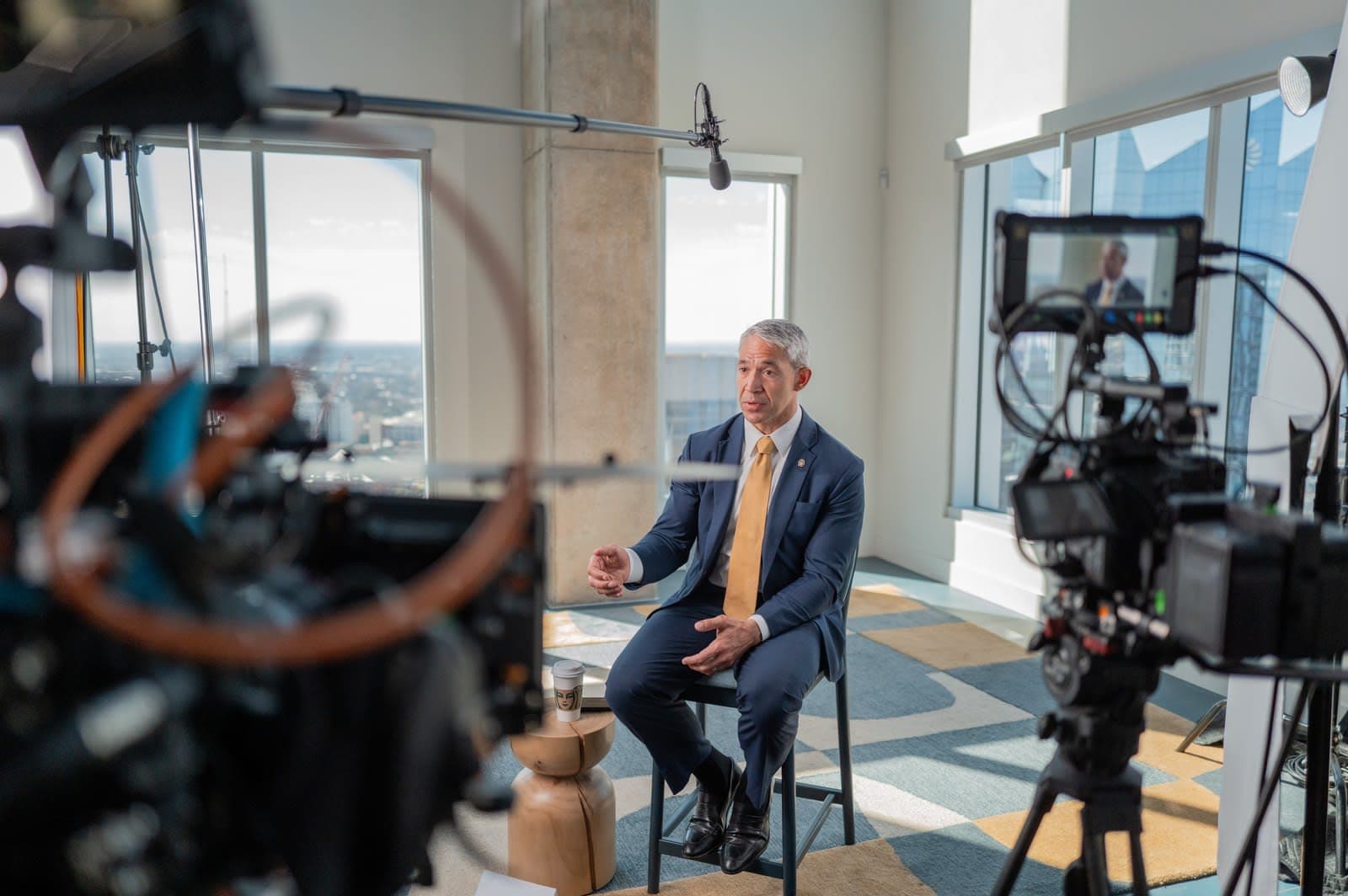 A two-camera crew interviews former San Antonio Mayor Ron Nirenberg by tall windows in a downtown high-rise.