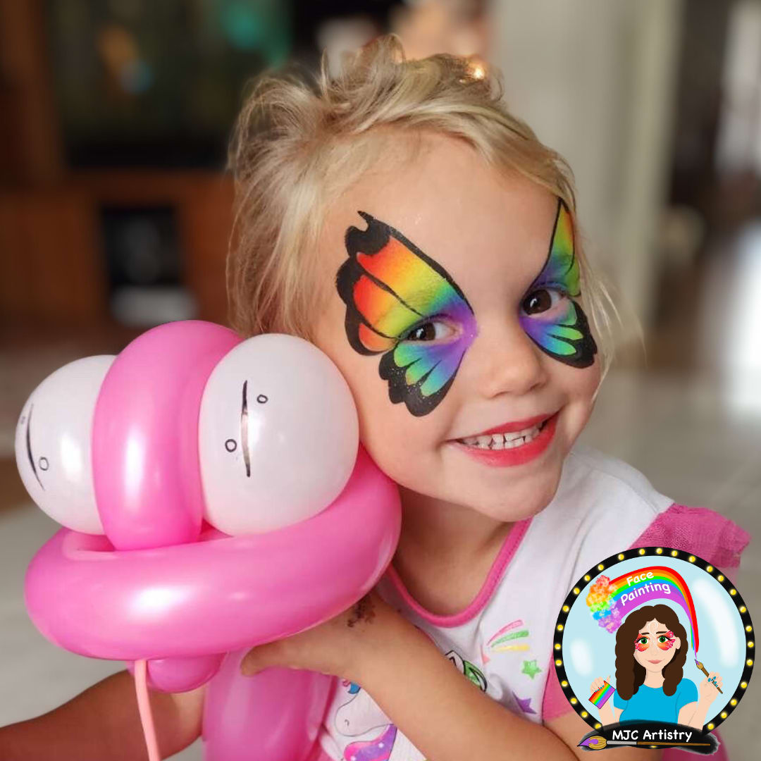 A blonde little girl with rainbow face paint holding a pink snake balloon at a birthday party in Coquitlam