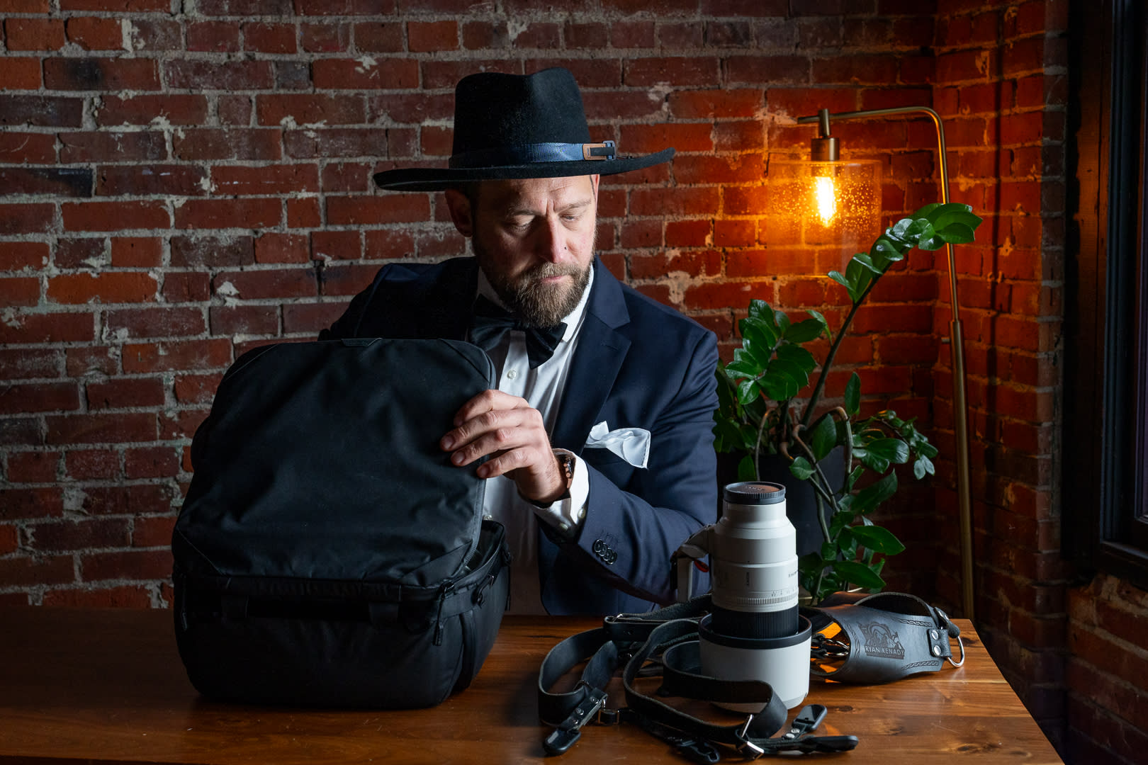 Photographer in a suit and hat preparing camera gear at a wooden table, with a camera bag and lens in a brick-walled studio s