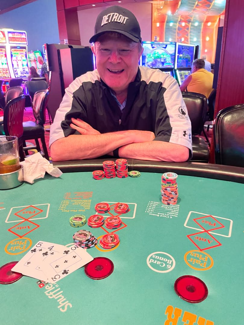 Man at Detroit cap sitting at casino poker table with cards and chips, four consecutive club cards visible on green felt.