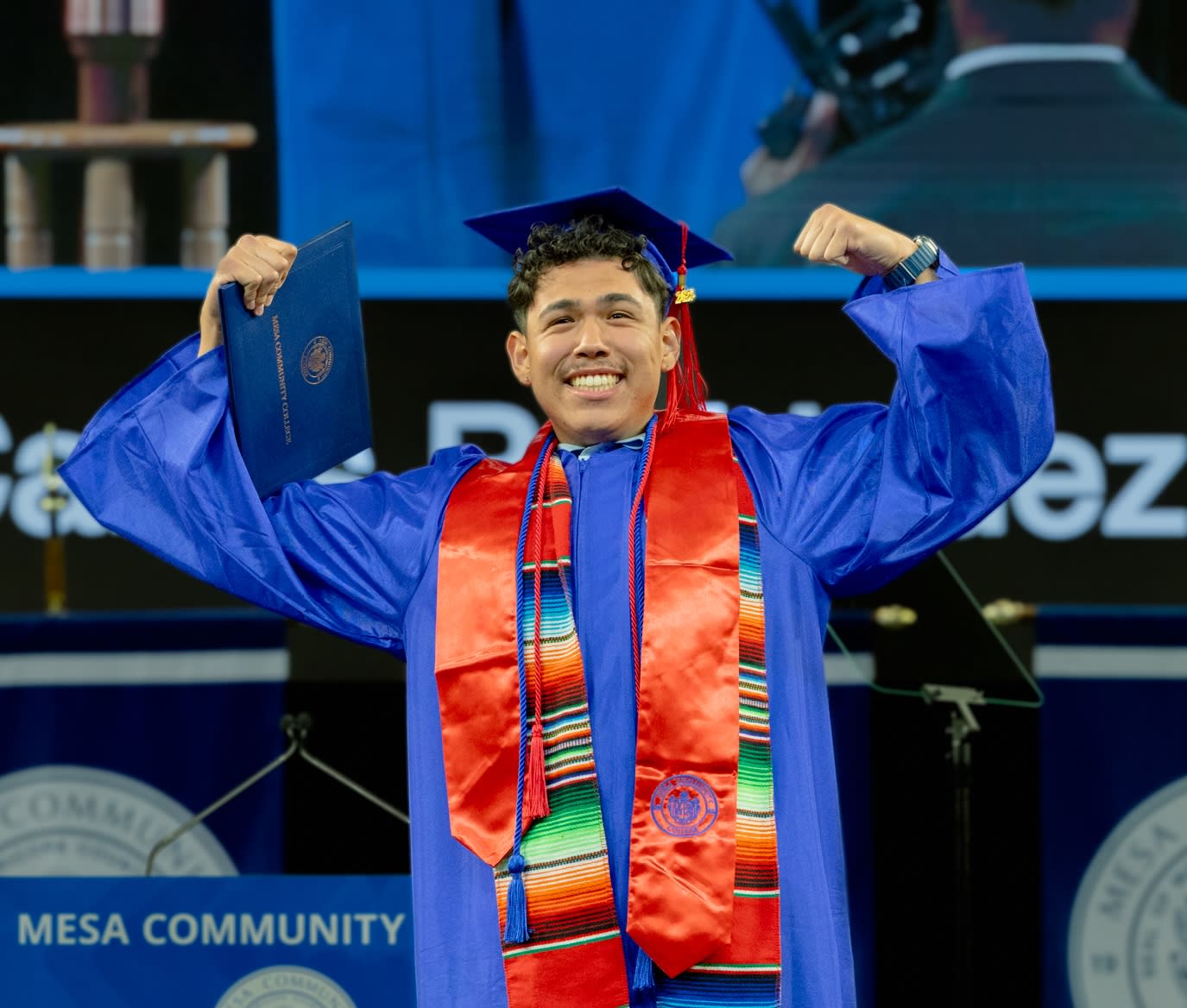 Man wearing a blue graduation cap and gown , posing with arms flexed