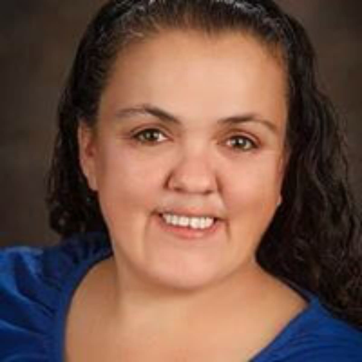 Headshot of woman with dark curly hair pulled back, bright blue top, friendly expression.