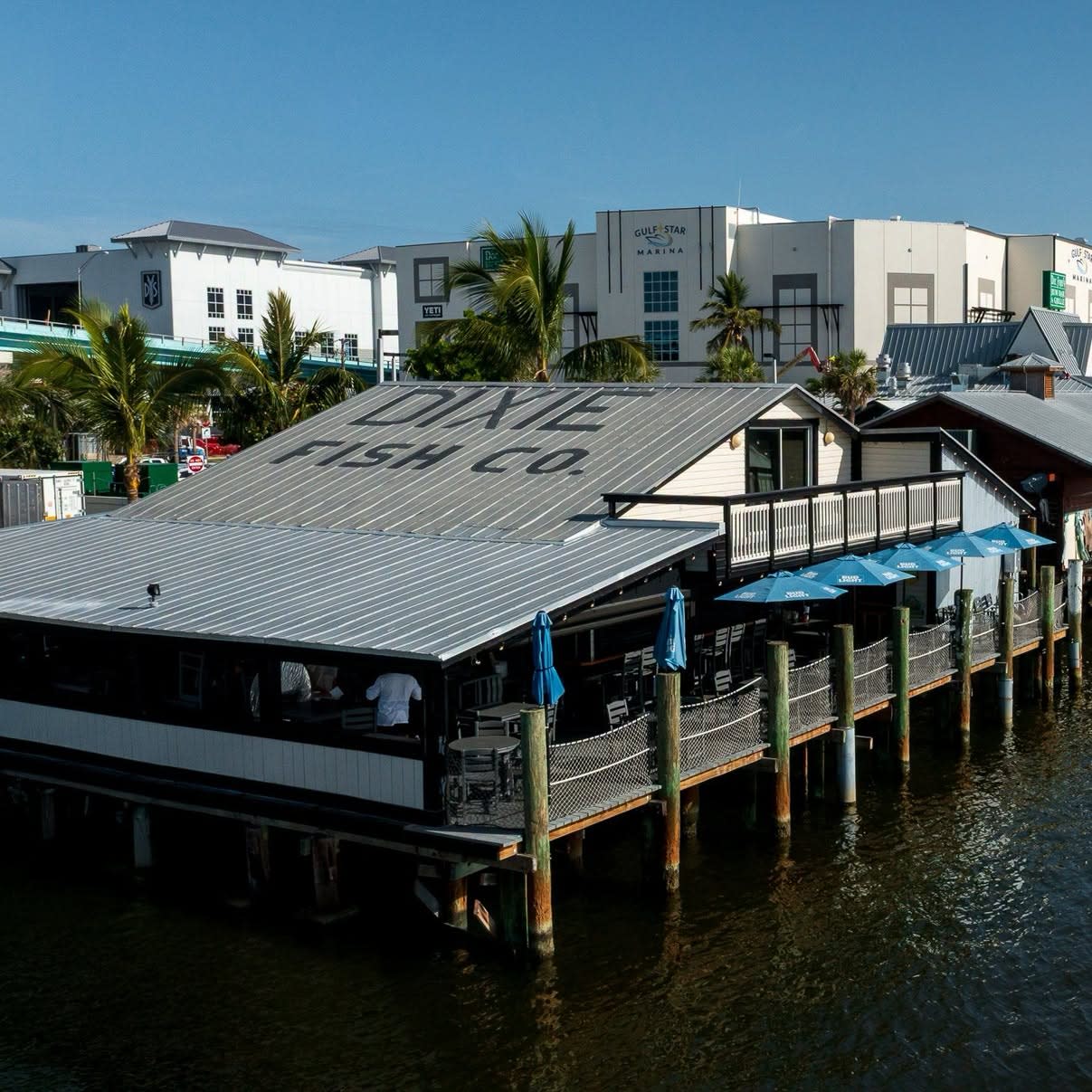 David Fish Co waterfront restaurant on wooden pilings with blue patio umbrellas, palm trees, and marina view