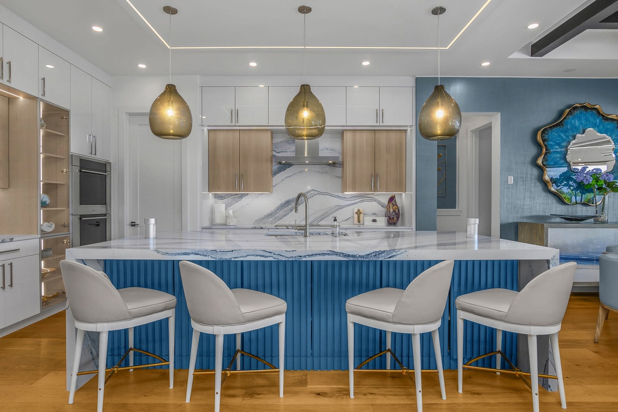 Modern kitchen with blue fluted island, white marble counter, four white stools, gold pendants, marble backsplash