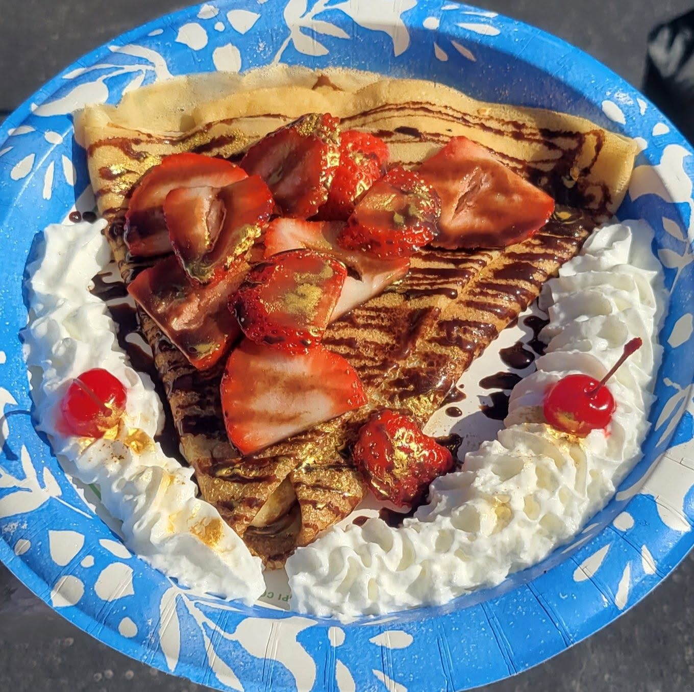 Sweet crepe topped with sliced strawberries, chocolate sauce, whipped cream, and maraschino cherries on blue plate.