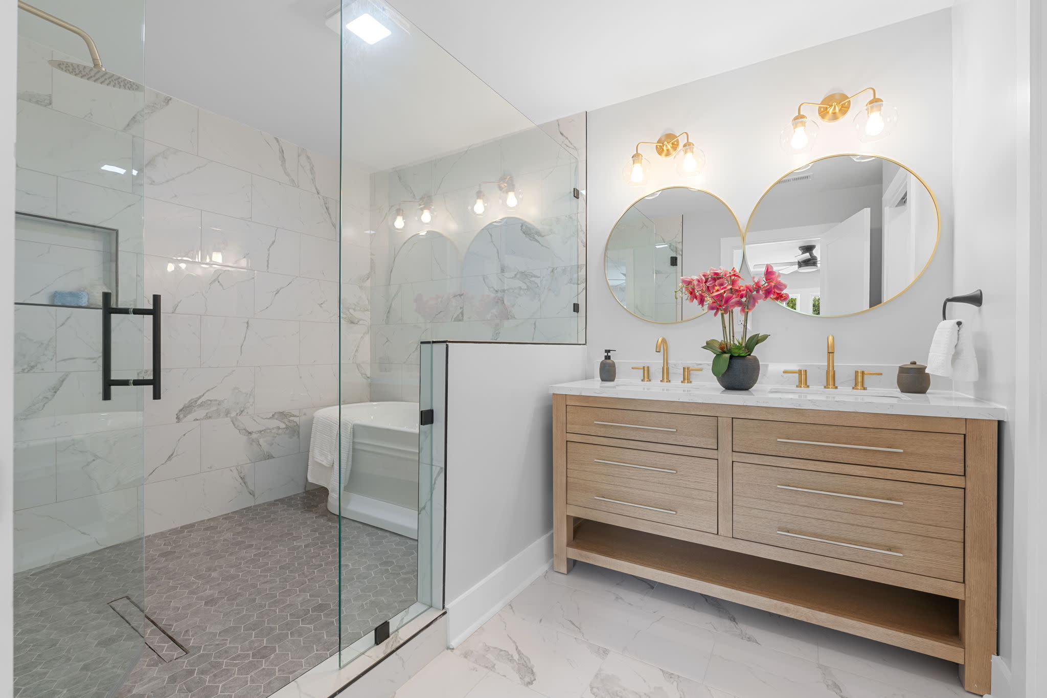 Luxury bathroom featuring a spacious glass enclosed shower, freestanding wood vanity, dual sinks, gold fixtures, and mirror