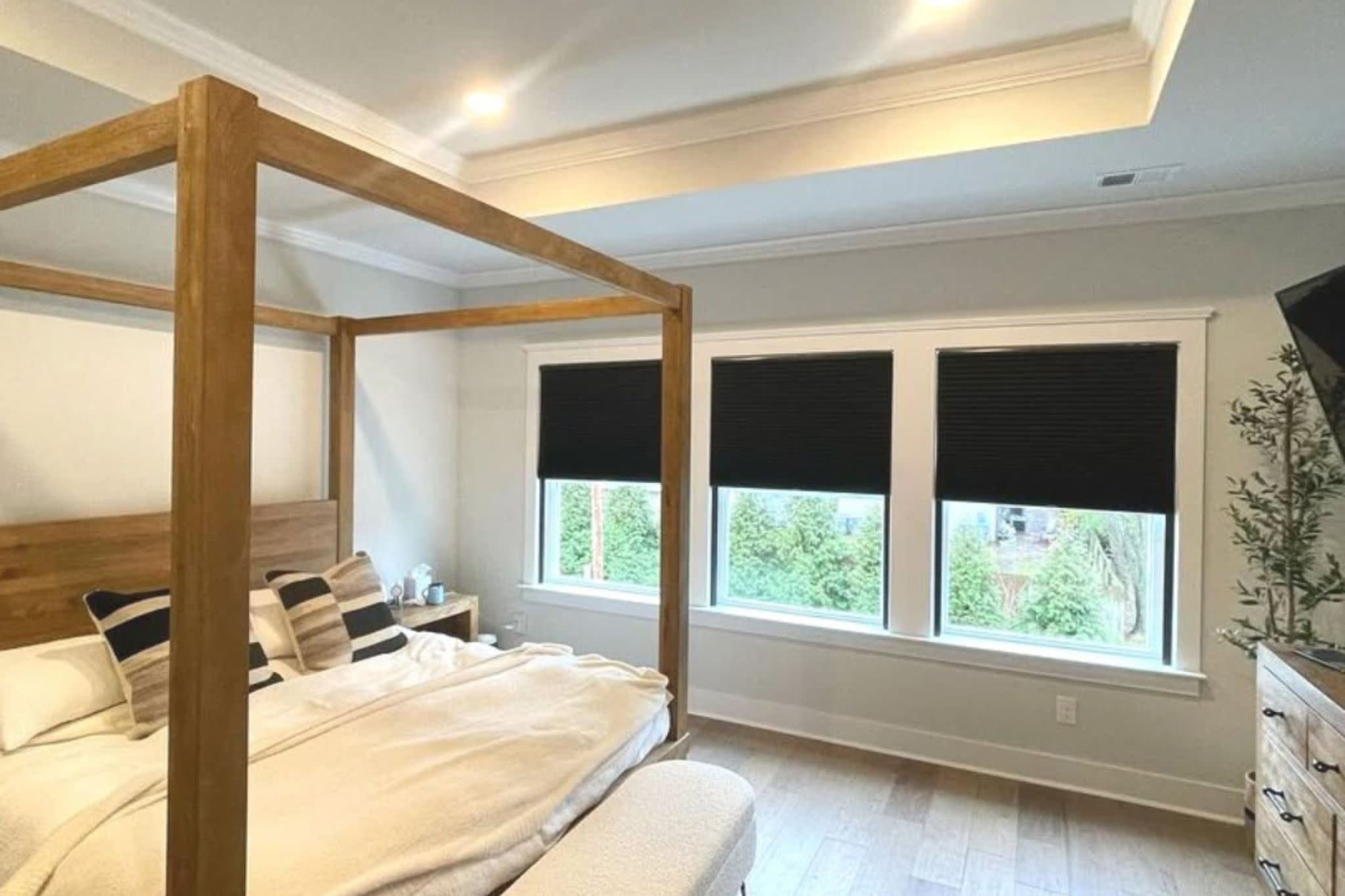 Dark blackout cellular shades partially lowered on three bedroom windows beside a wooden canopy bed with cream bedding.