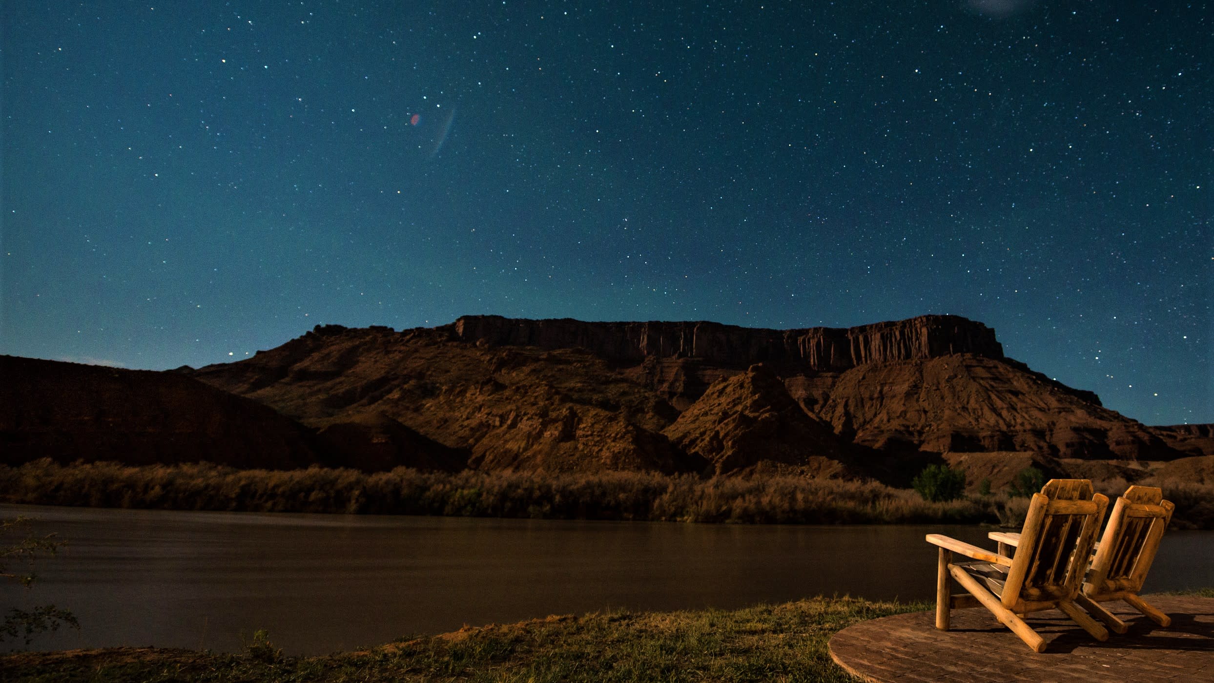 Two wooden lounge chairs overlook the Colorado River beneath a star-filled sky at Sorrel River Ranch, with dramatic red rock