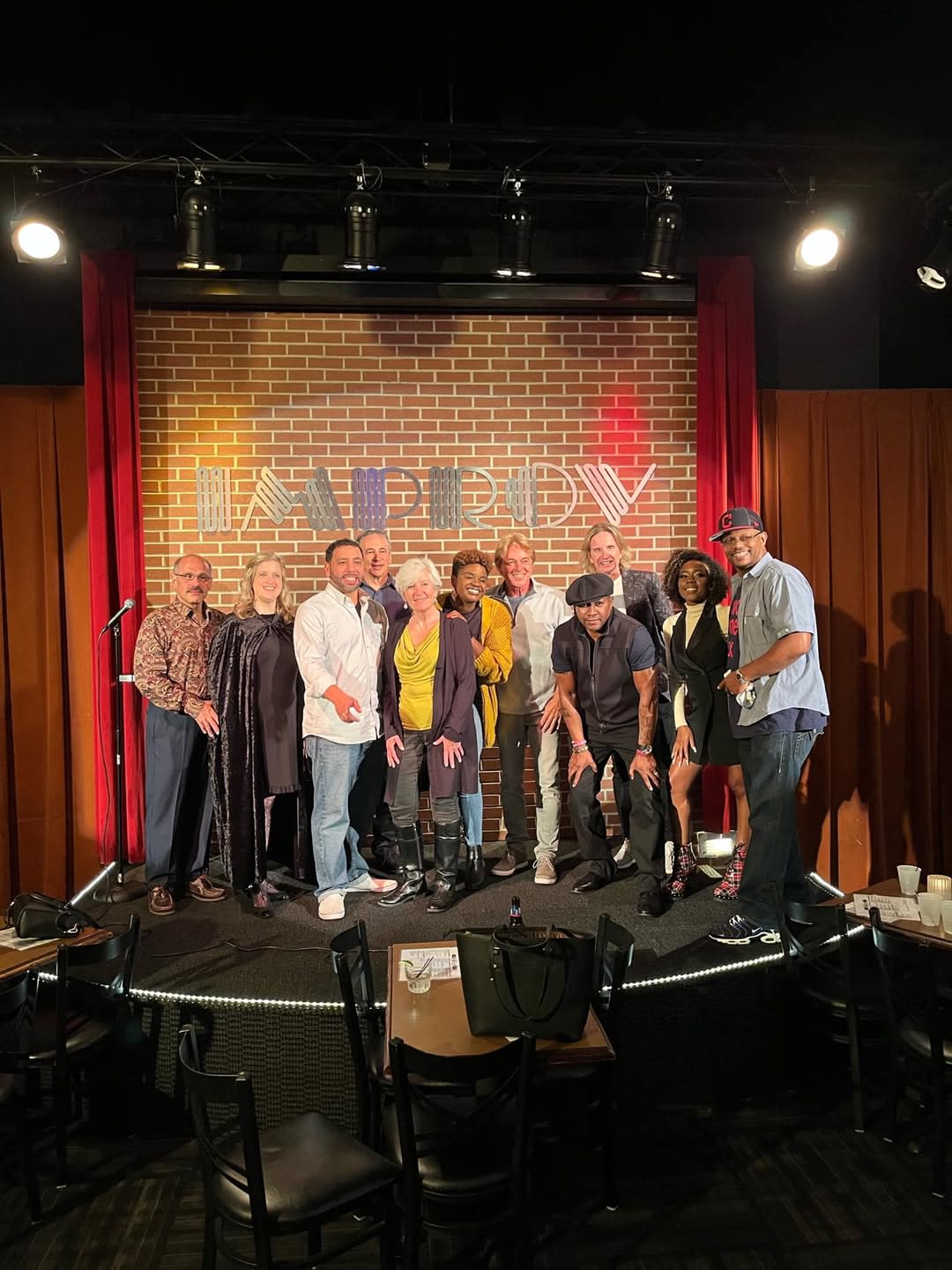 Ten people smile and pose together on a comedy club stage beneath an “IMPROV” sign, with a brick backdrop, red curtains, and