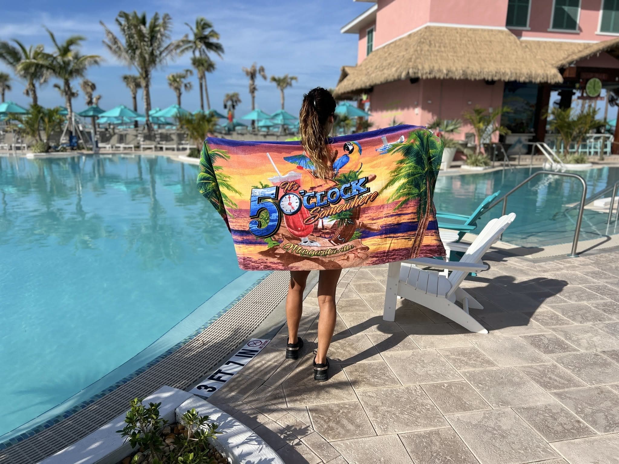A woman stands by a pool holding a colorful '5 O'CLOCK' beach towel with palm trees and a parrot.