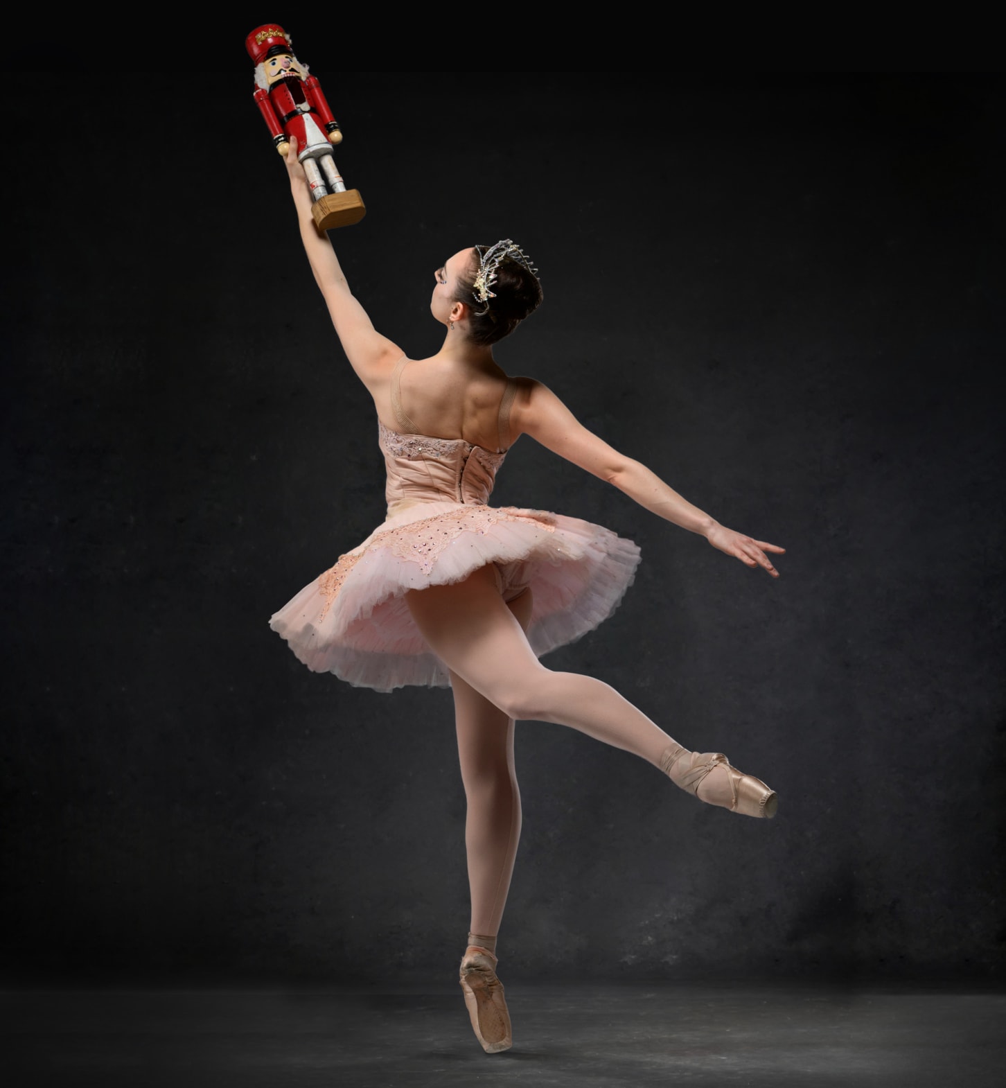 A ballerina en pointe in a pink tutu and crown holding up a wooden nutcracker