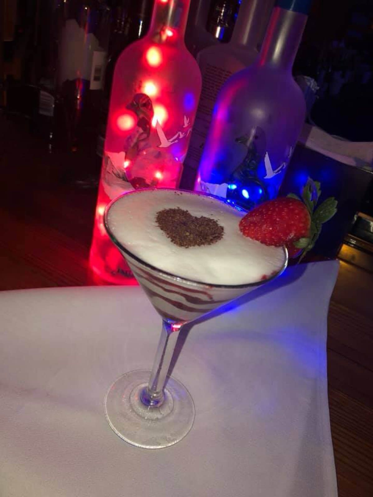 Martini glass with strawberry and heart-shaped topping, colorful LED-lit bottles behind on bar
