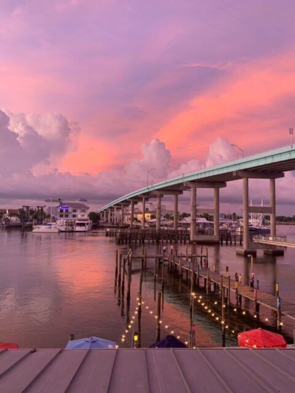 Sunset over marina with bridge spanning calm water, pink-purple sky reflecting on surface.