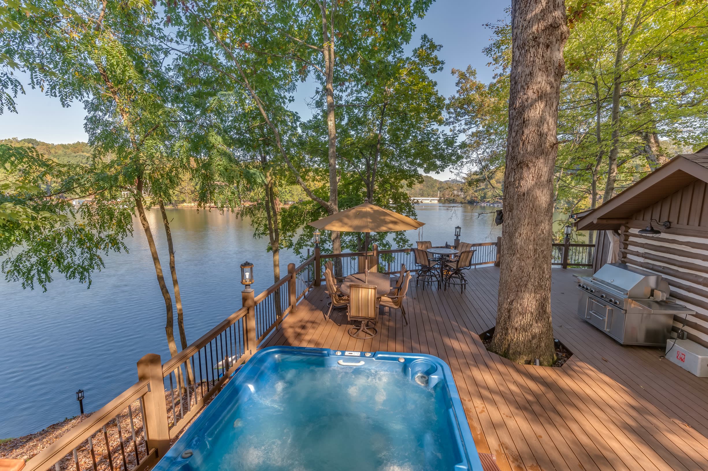 Lakefront deck with a private hot tub, outdoor dining area, gas grill, and peaceful waterfront views surrounded by mature tre