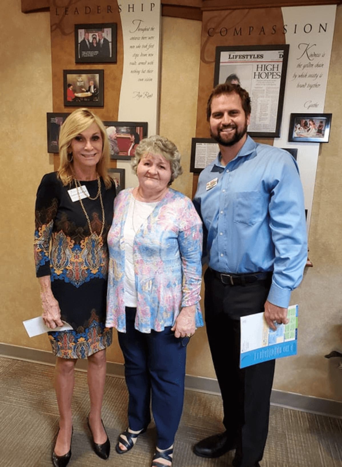 Three people posing together in office hallway with framed photos and inspirational words on walls.