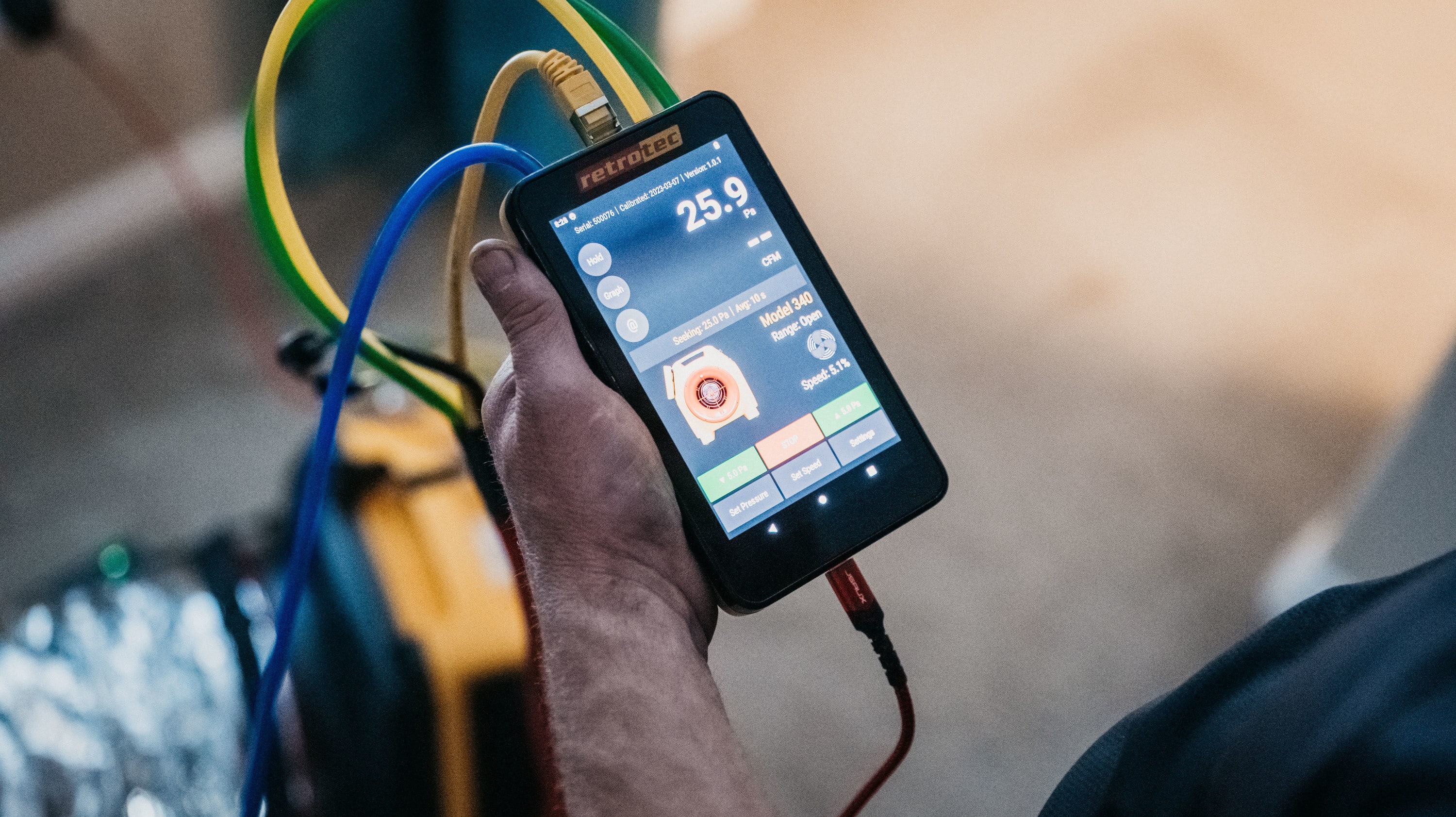 Close-up of a hand holding a Retrotec digital device with colorful cables connected, measuring pressure in HVAC testing.