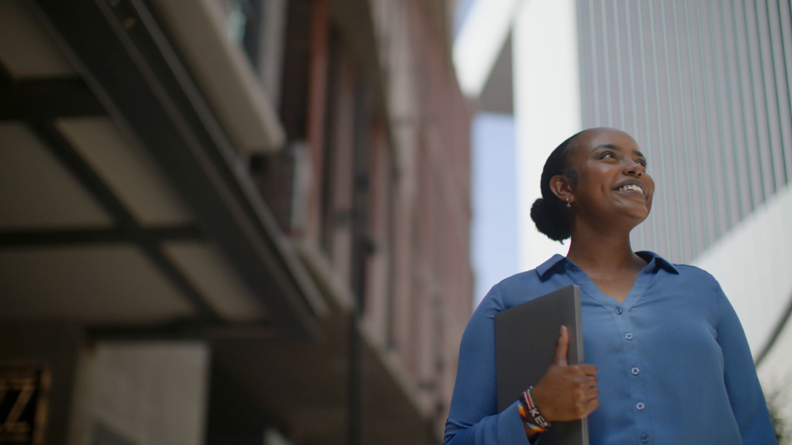 NPower Dallas alum Valentine Komen, holding a laptop, smiles as she looks up between city buildings.