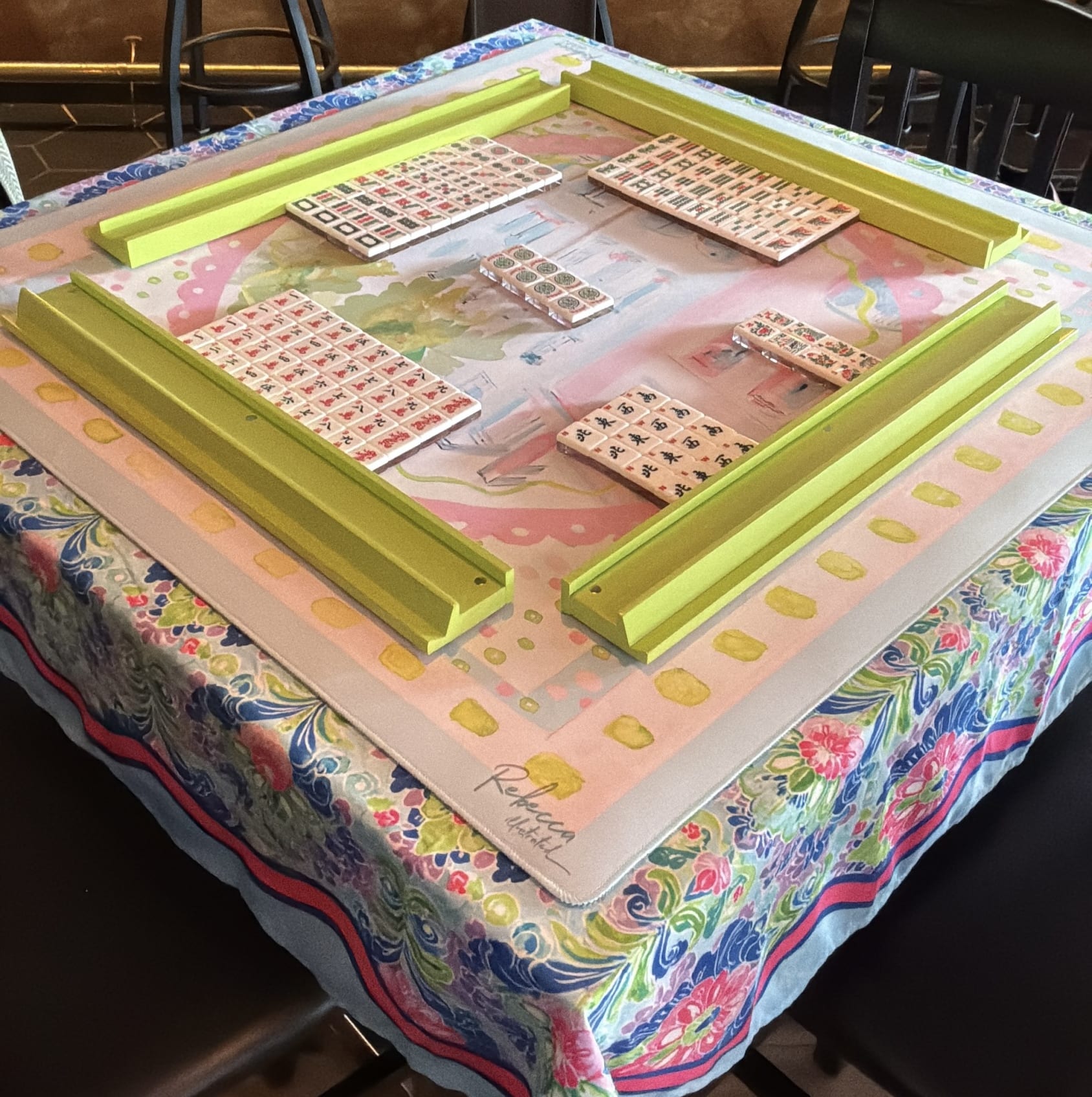 Mahjong tiles separated into suits and bordered by lime green racks on a colorful blue, pink and green floral tablecloth