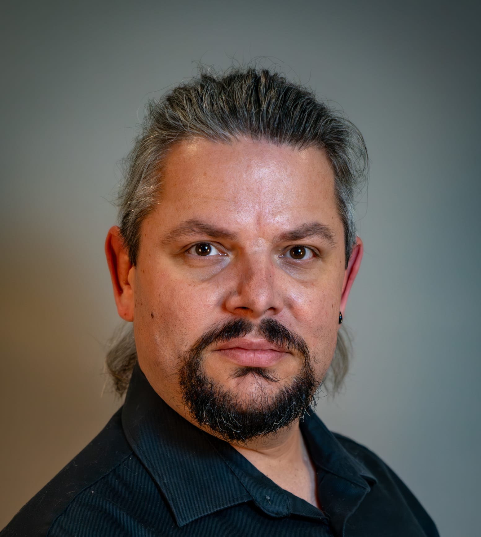 A Man wearing a black button up in front of a dark white background posing for the camera