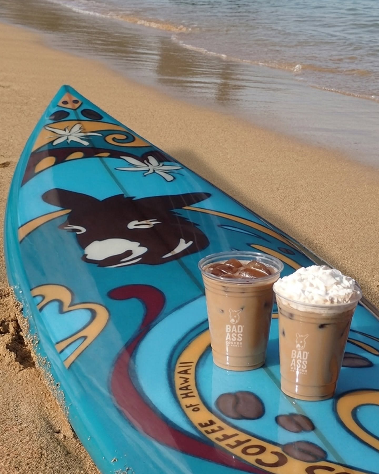 Blue paddleboard with Bad Ass Coffee drinks resting on sandy beach near gentle ocean waves