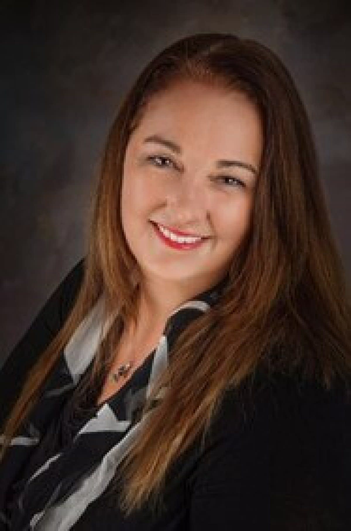 Smiling woman with long brown hair, wearing black blazer and striped blouse, studio portrait.