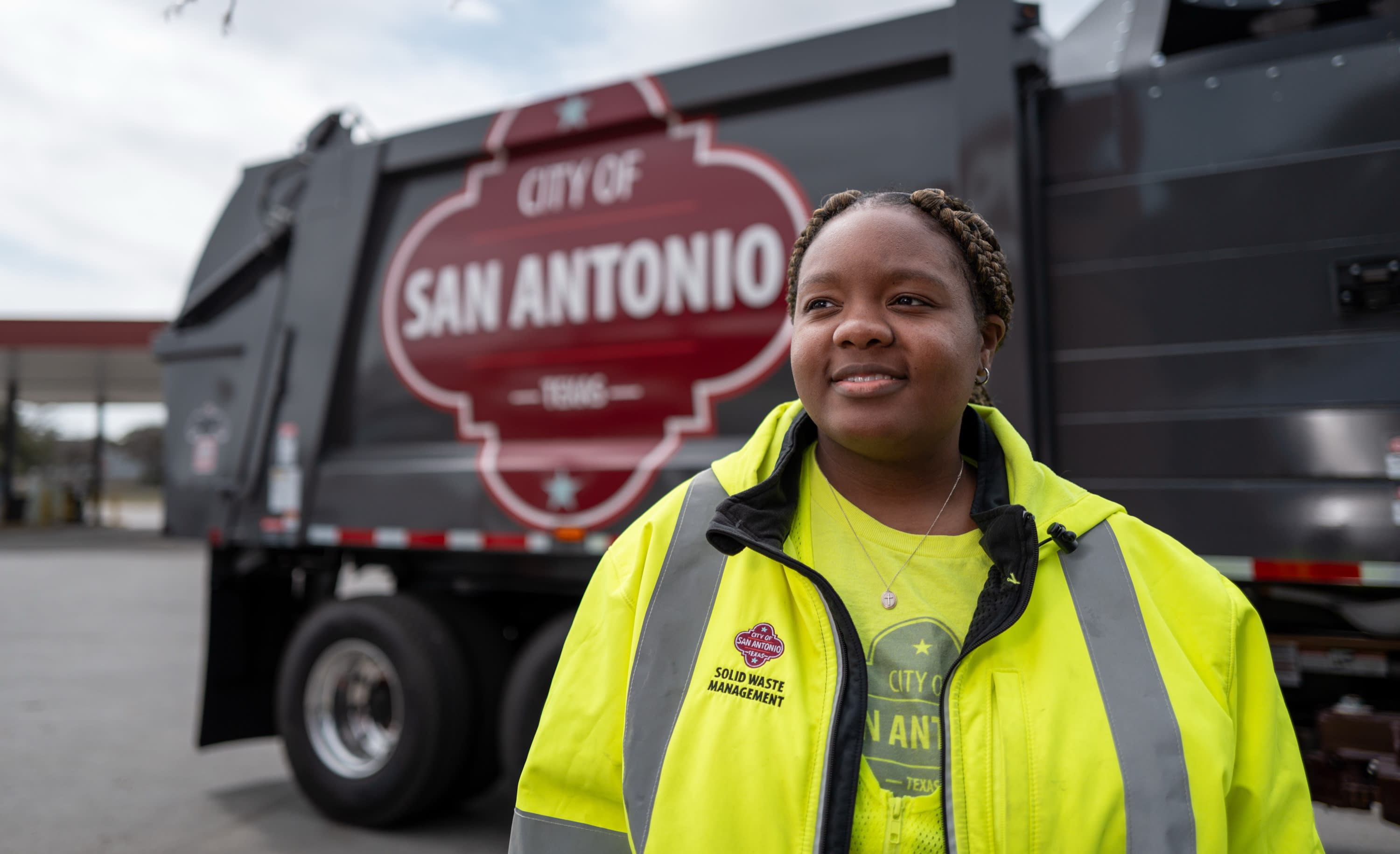A City of San Antonio Solid Waste Management CDL apprentice in a yellow safety jacket smiles in front of a city truck.