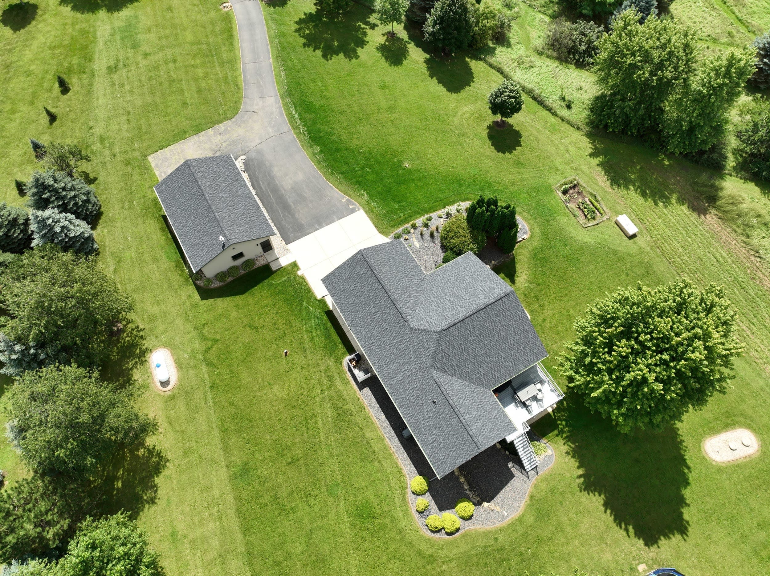 Drone photograph of residential home and garage covered in dark shingles, surrounded by trees and greeney