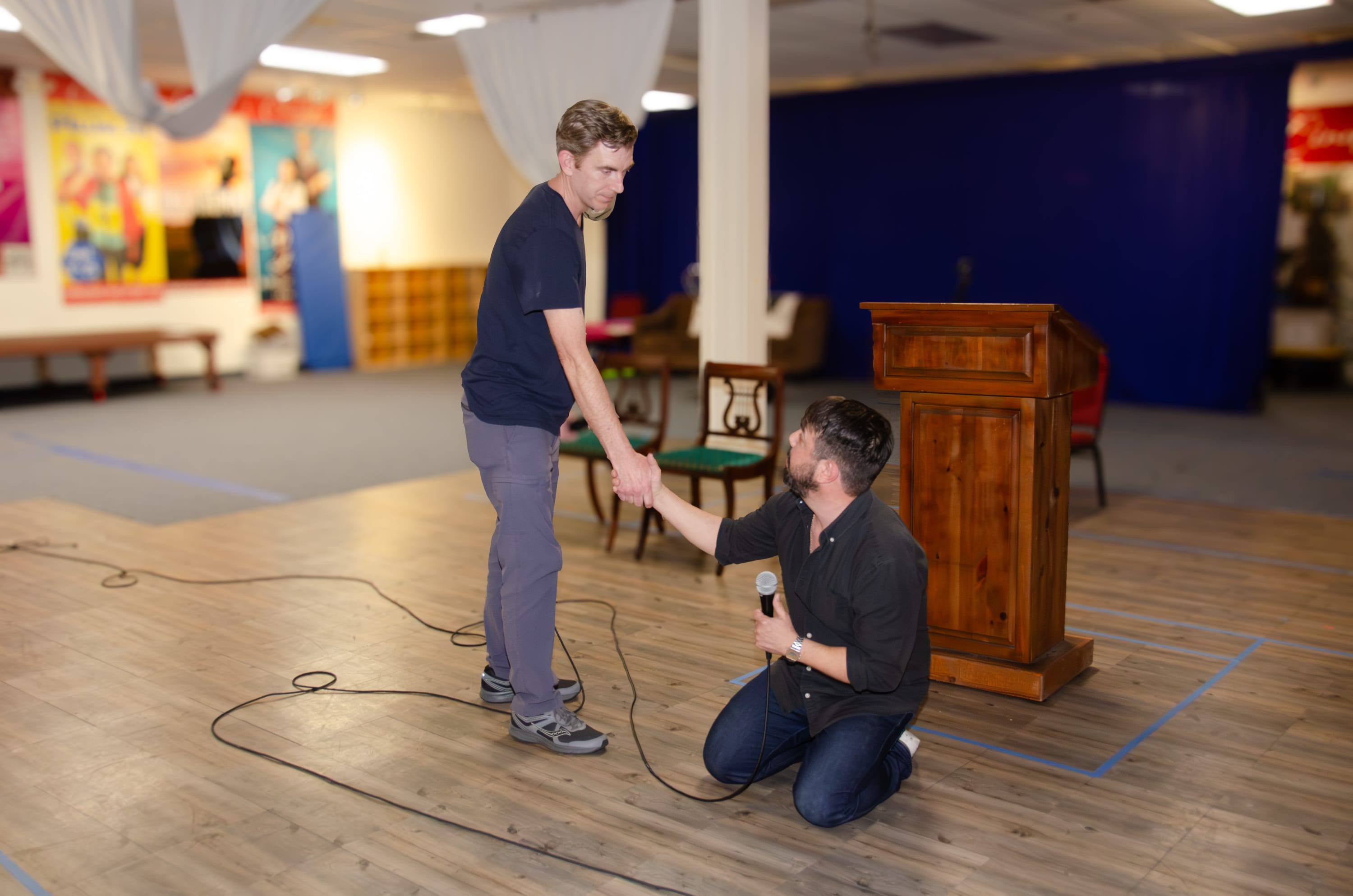 Two men on a rehearsal stage share a serious moment, with one kneeling and holding a microphone while reaching up to clasp th