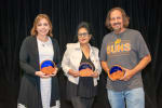 Three adults hold tan and blue awards as they stand in a group.
