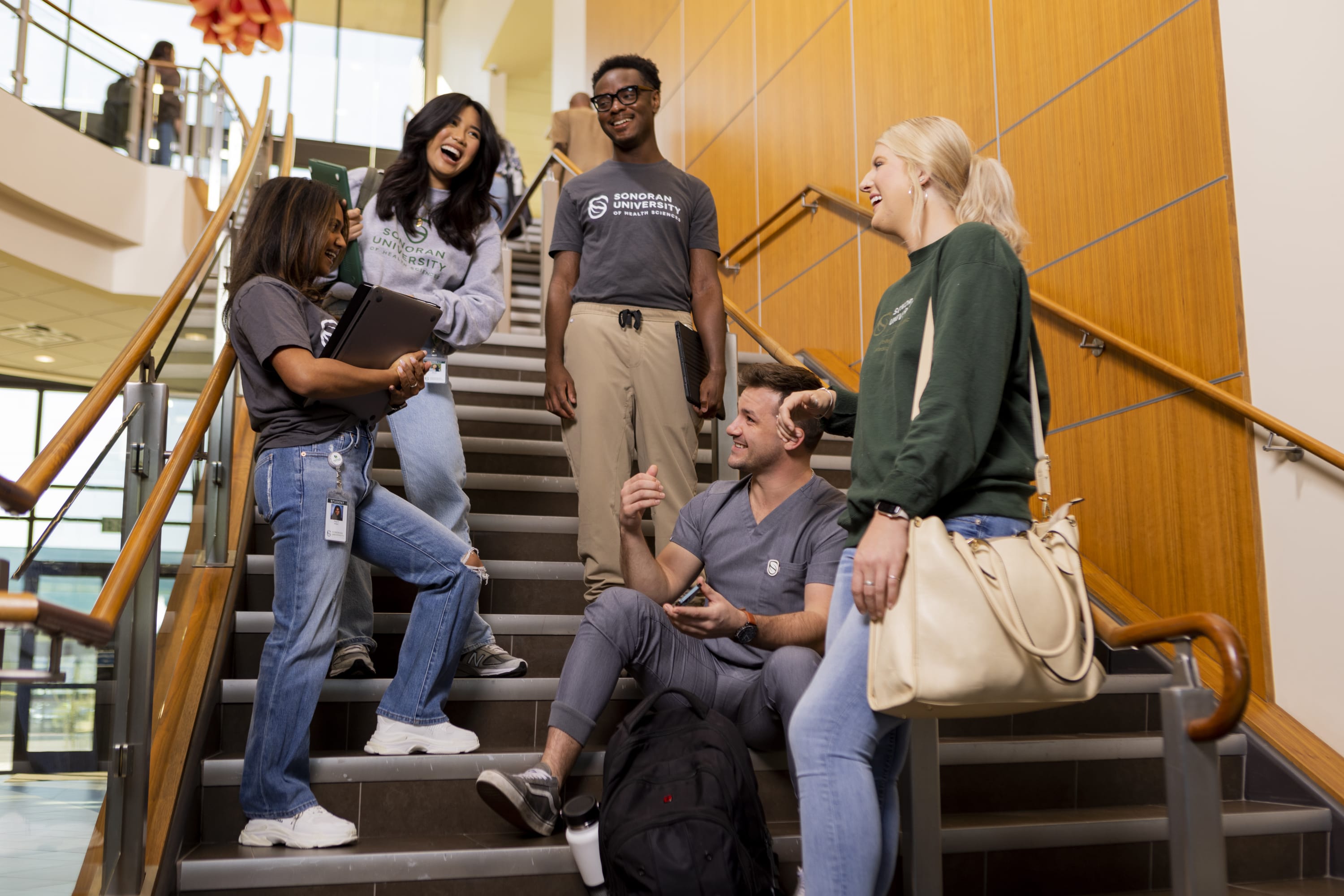 A group of students gather on an indoor stairway, talking and holding notebooks and a backpack inside a university building.