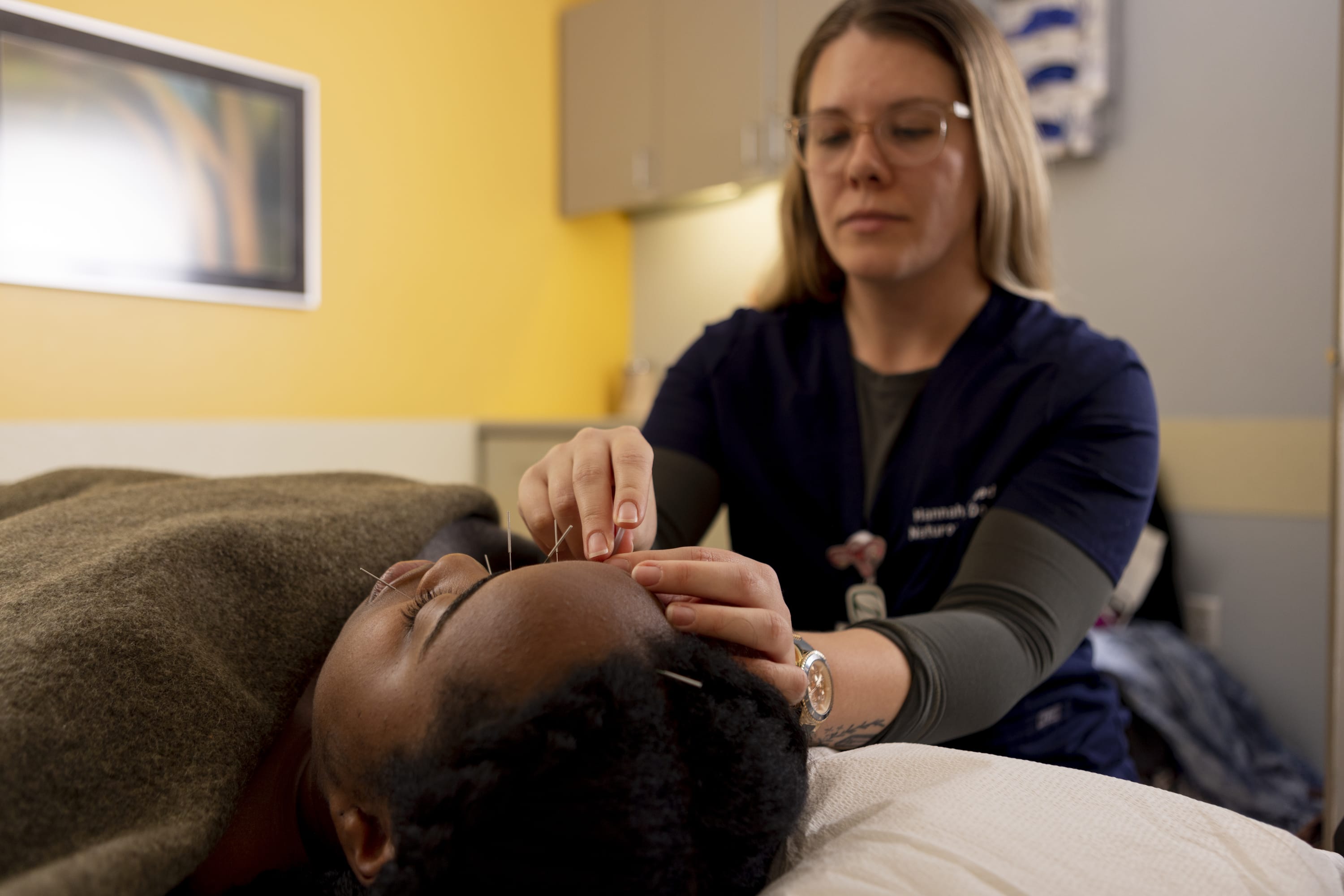 A clinical student performs acupuncture on a patient lying on a treatment table in a clinical exam room.