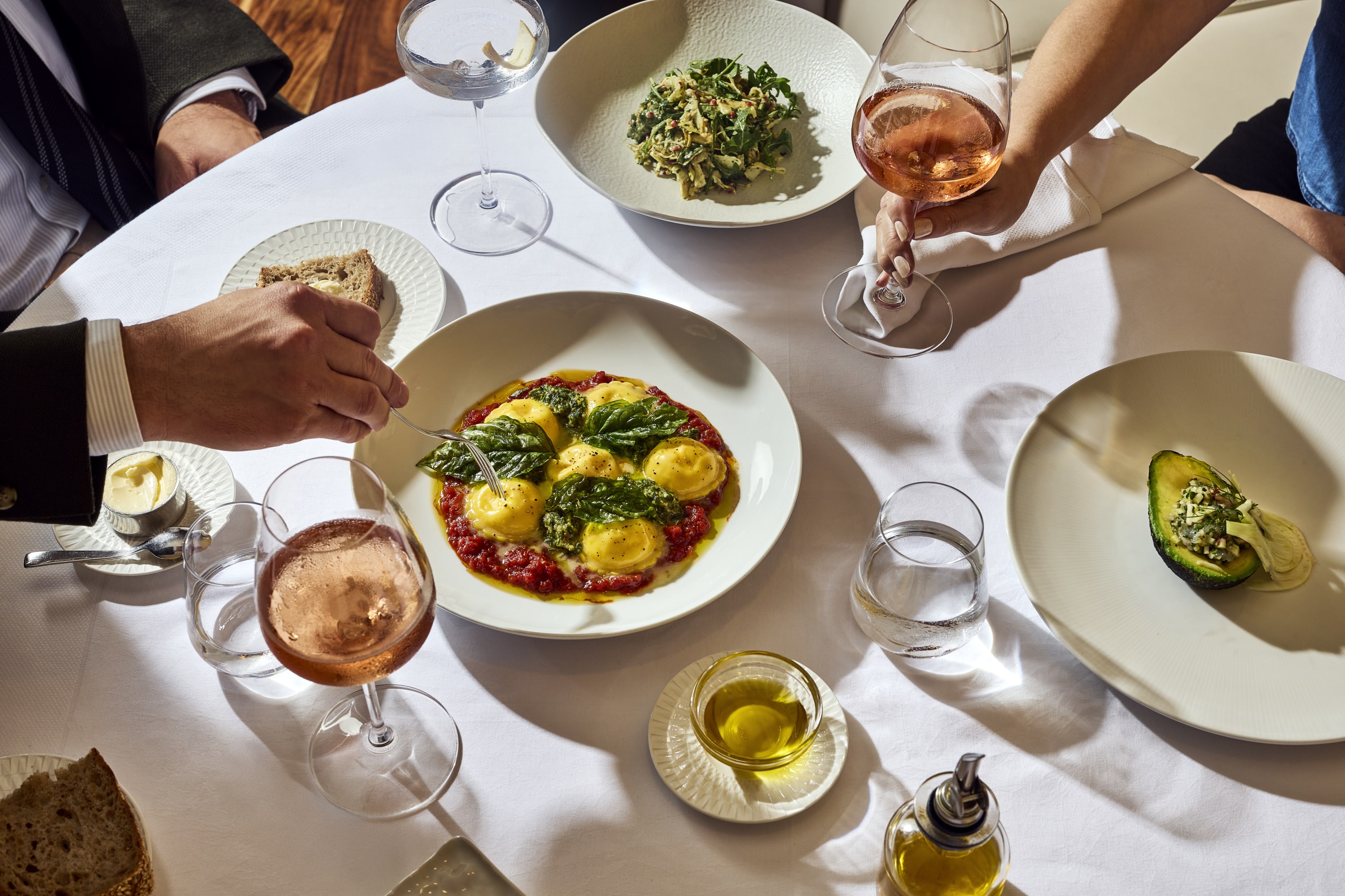 Overhead view of a sunlit restaurant table with pasta, avocado, bread, olive oil, water, and rosé glasses being enjoyed durin