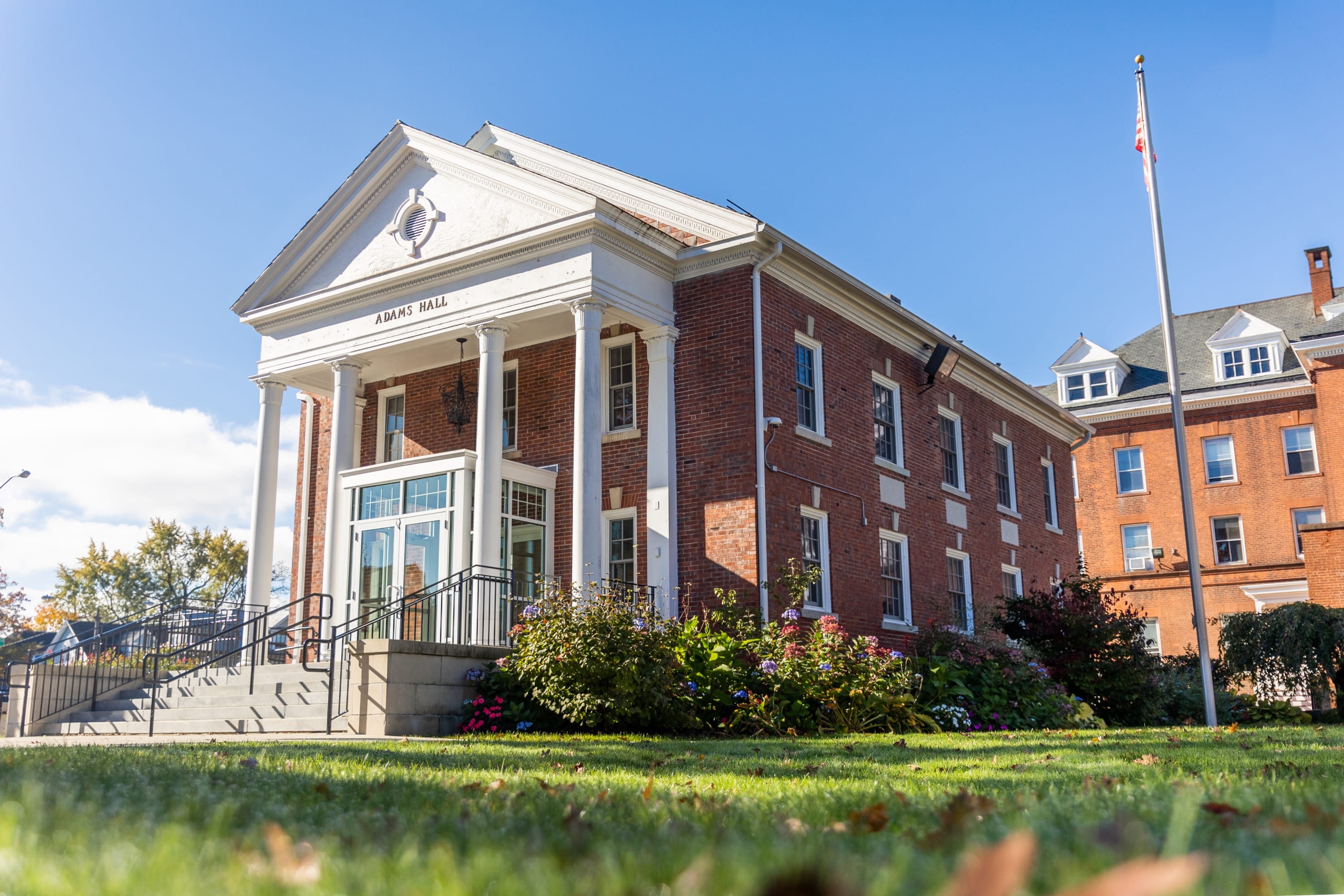 Exterior view of AIC’s Adams Hall with white columns, brick facade, landscaped lawn, and an American flag on a sunny day