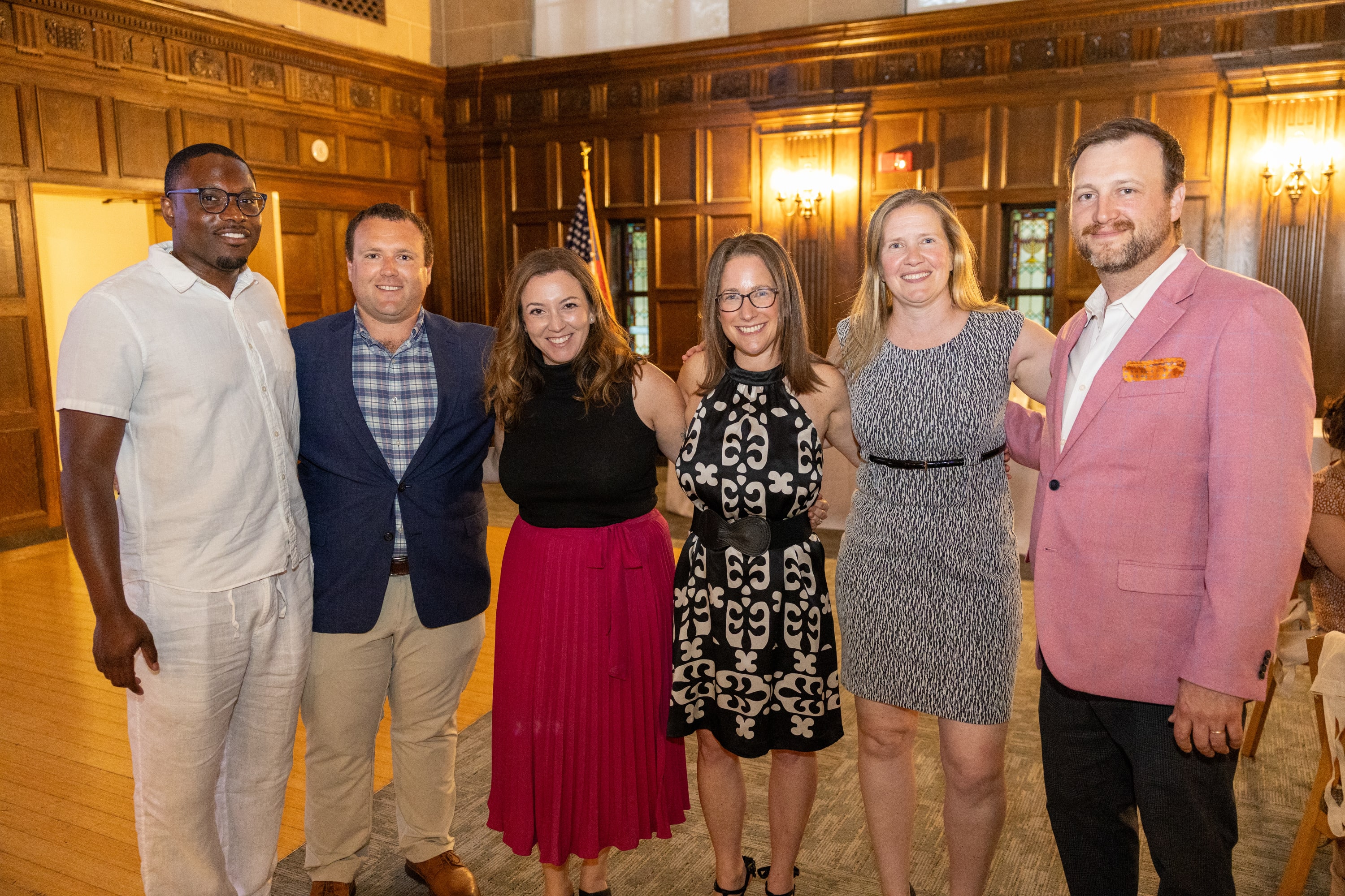 Six people posing arm-in-arm for a group photo in a wood-paneled event hall.