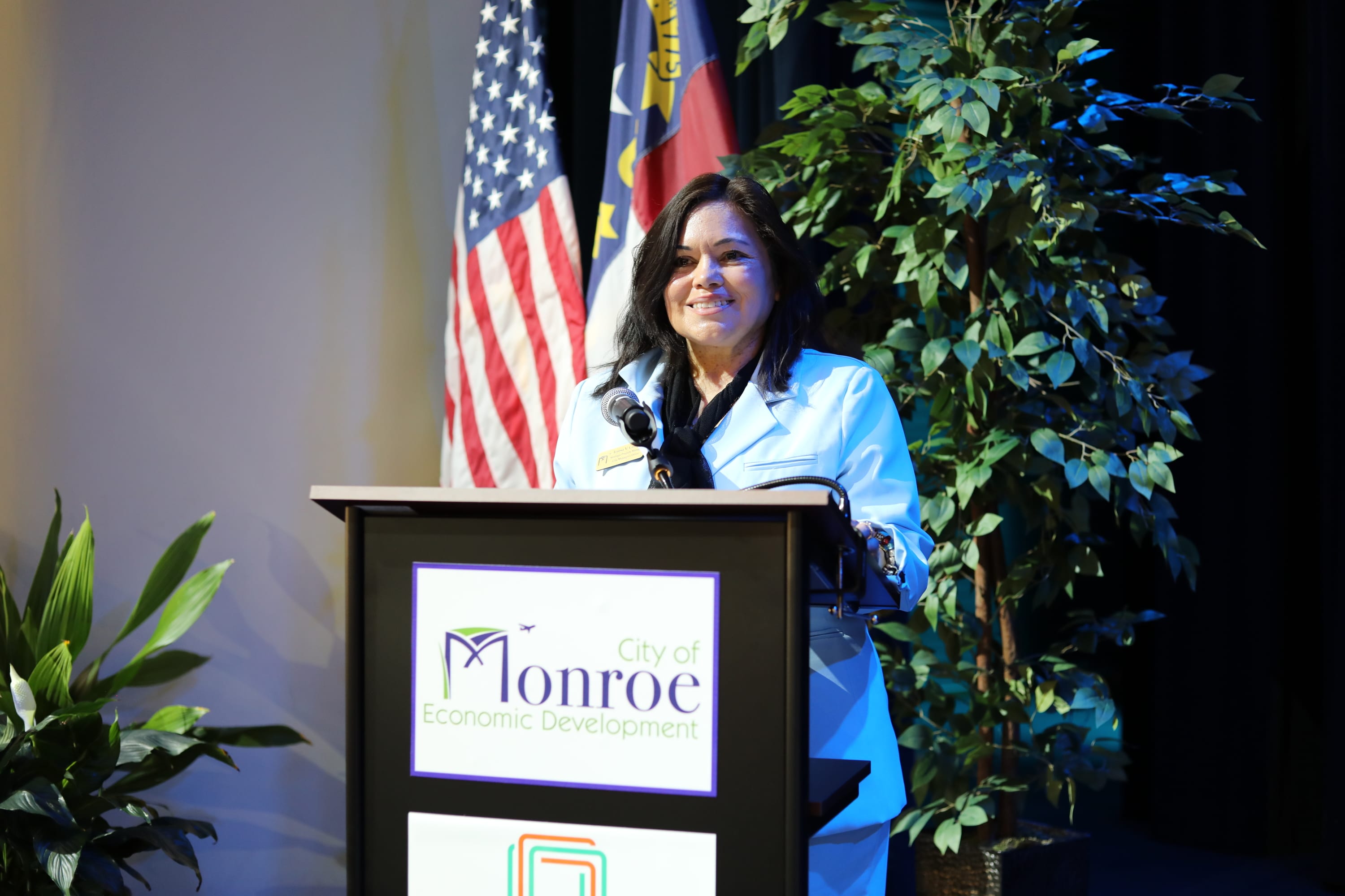 Teresa Campo Economic Development Director Photo Petite Woman Olive Toned Dark Hair Speaking at a podium with USA and NC Flag