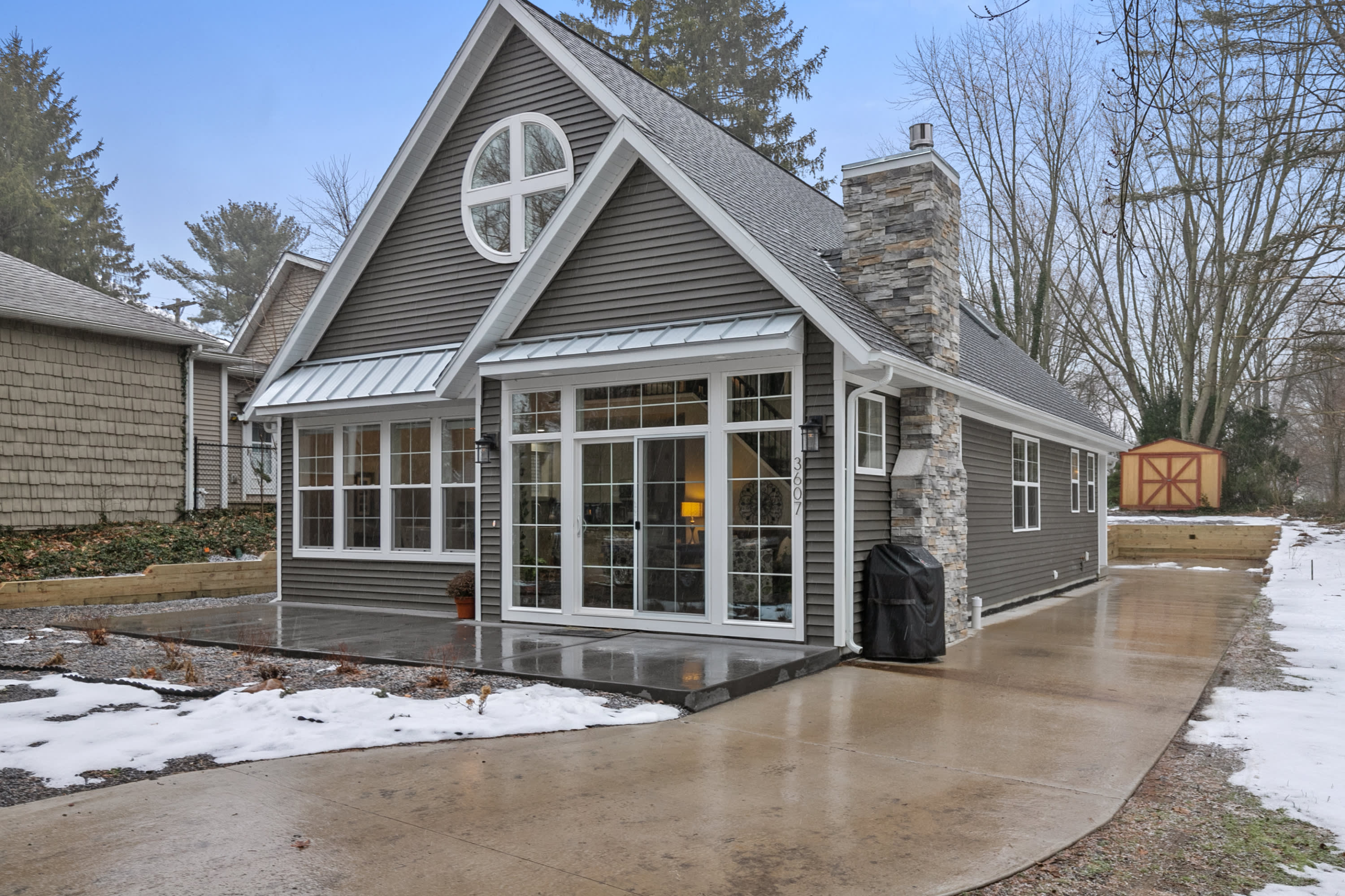 Cottage-style custom home with gray siding, round gable window, stone chimney, and large windows by Spaces Designer Homes
