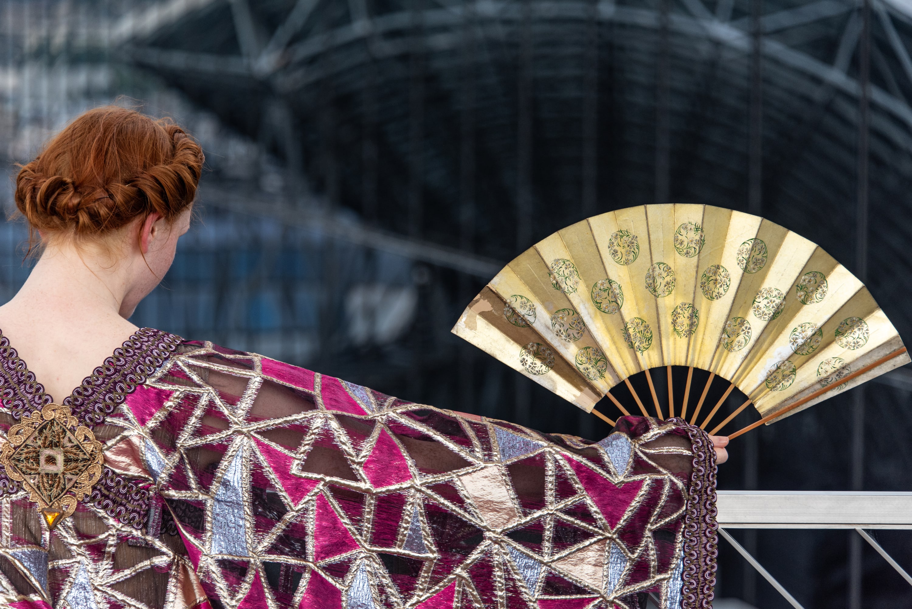 A performer in an ornate costume holds a gold folding fan against a modern architectural backdrop.