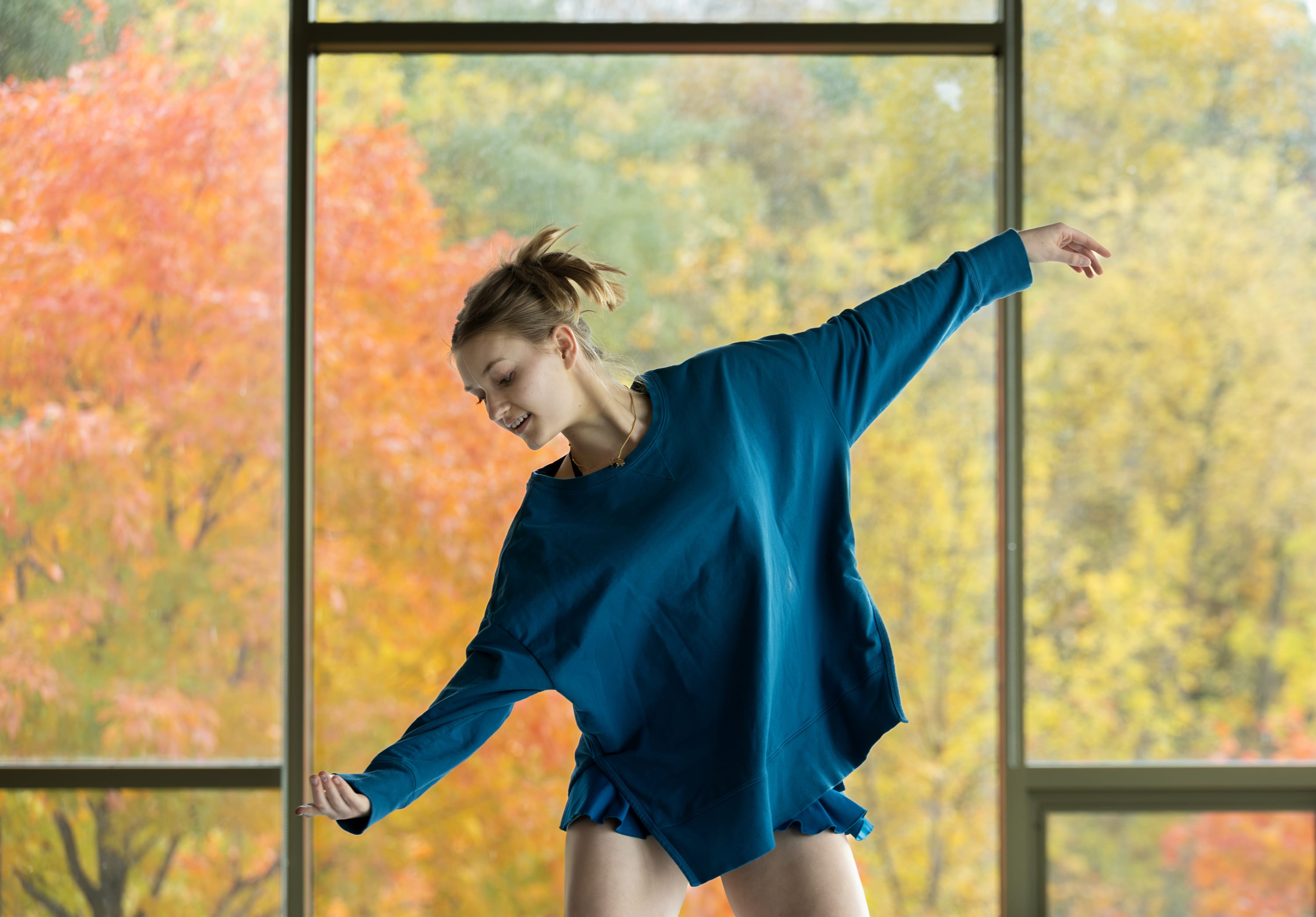 A dancer practices in front of large floor-to-ceiling windows, extending both arms while moving across a studio space.