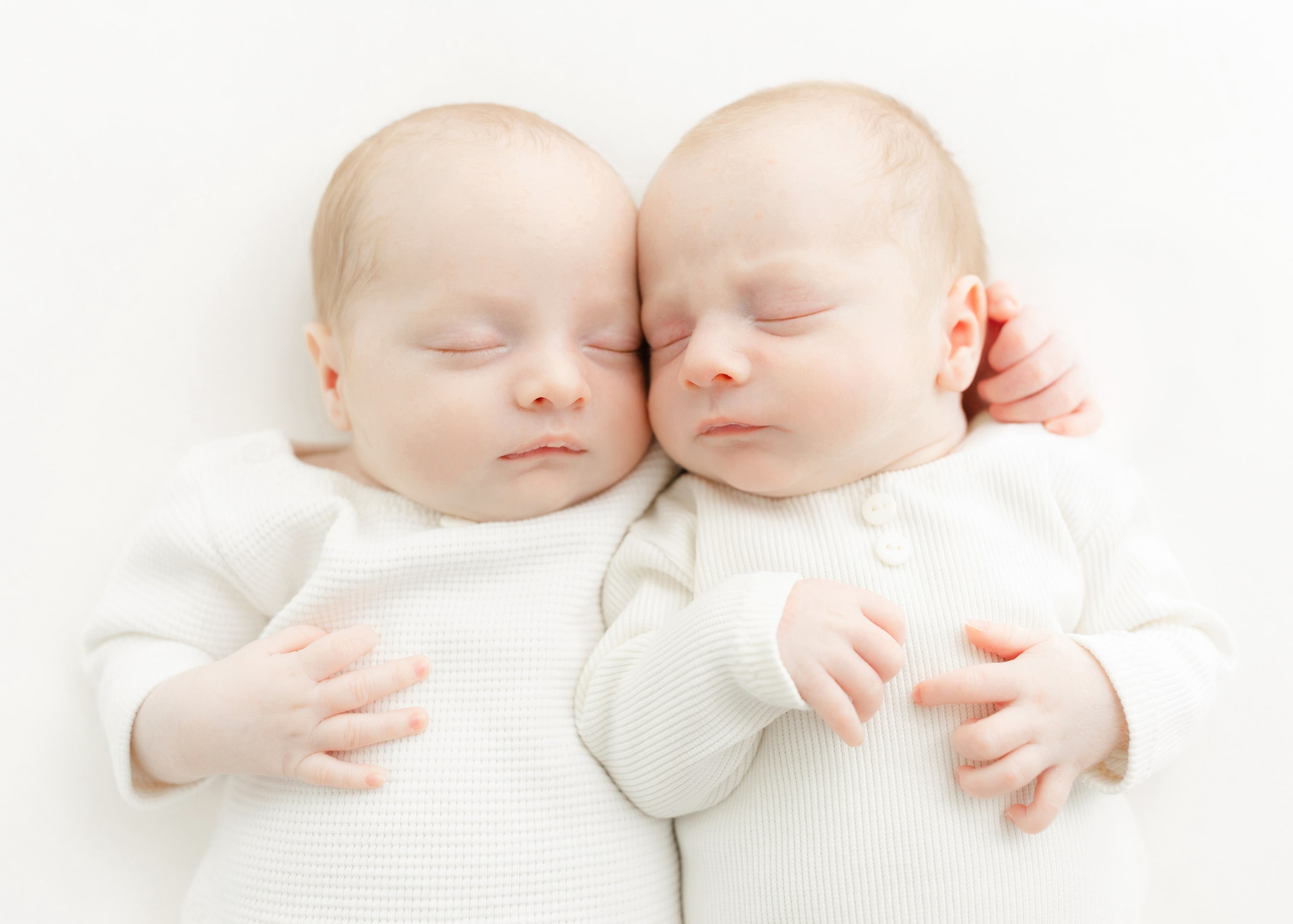 Sleeping newborn twin babies cuddled together on a white backdrop, wearing soft neutral outfits during a light and airy newbo