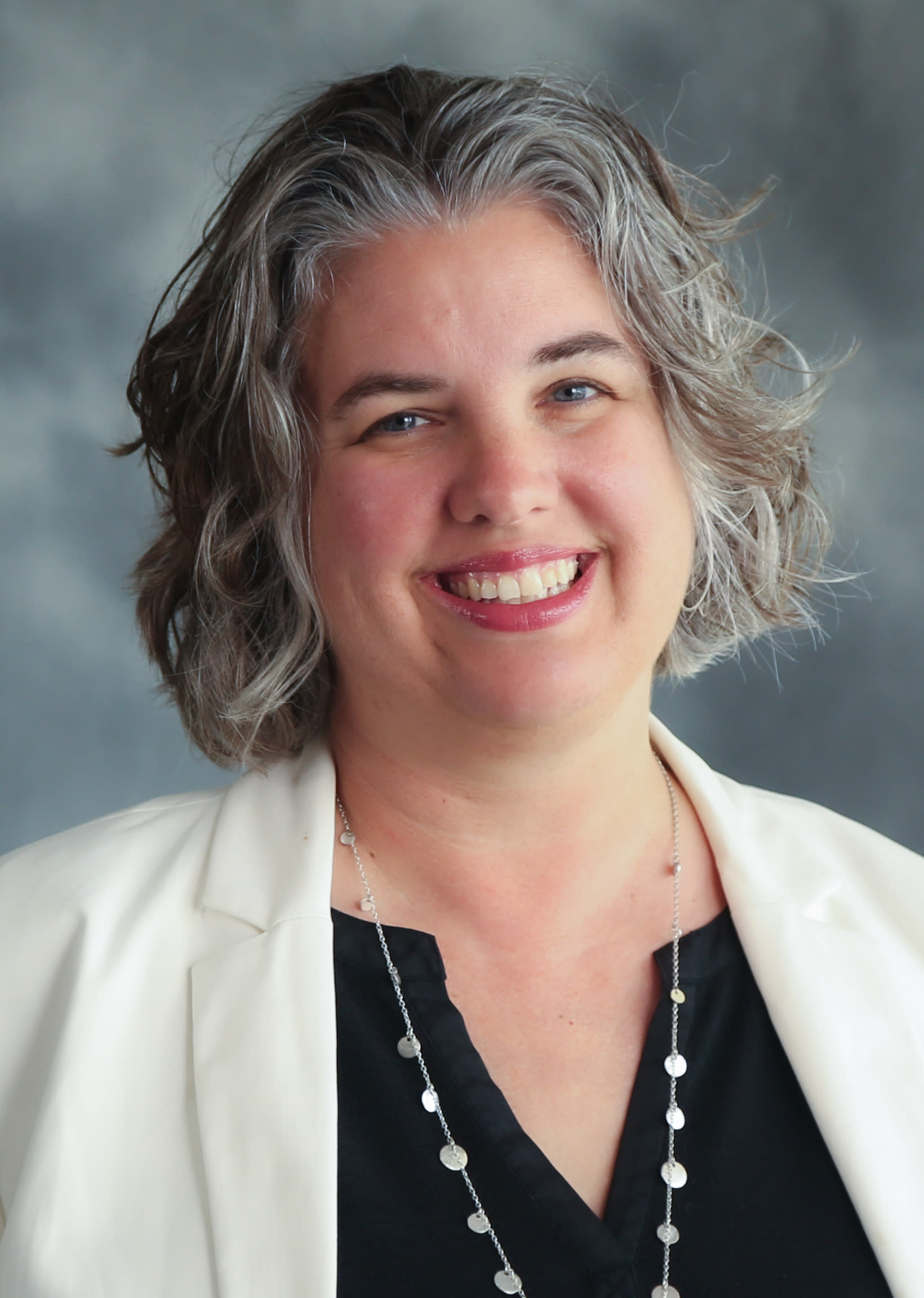 Marsha is wearing a white jacket over a black top and a long silver necklace.